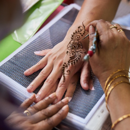 A person is applying detailed henna designs on another's hand, with floral and intricate patterns visible.