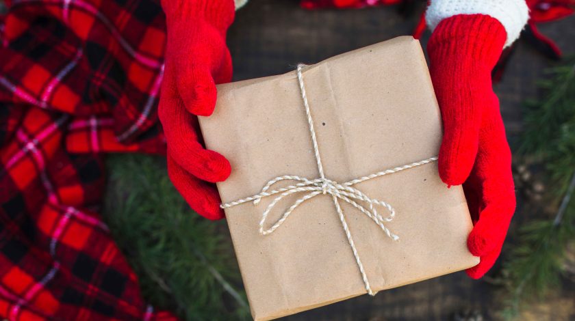 A person wearing bright red gloves holds a neatly wrapped brown paper gift tied with twine, set against a backdrop of festive red plaid fabric and green pine branches, creating a cozy holiday atmosphere.