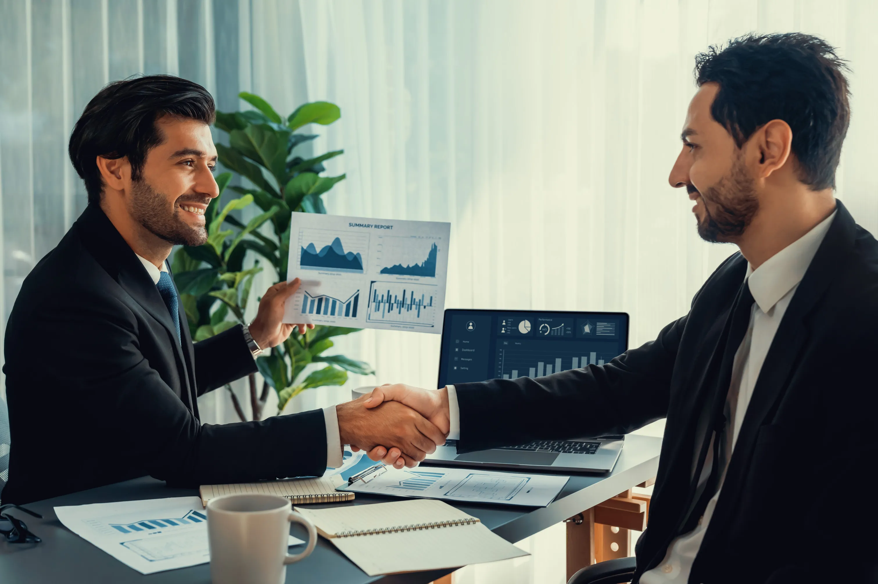 Two businessmen in suits shaking hands over a desk with charts, reports, and a laptop displaying data and graphs.