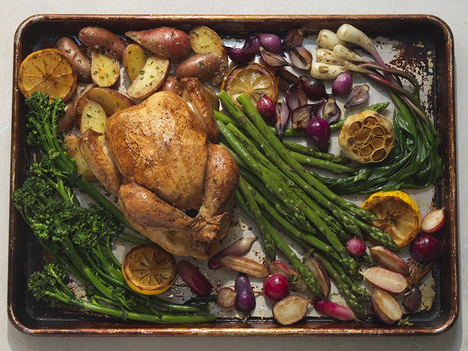 A whole golden brown roasted chicken on a sheet pan styled with broccolini, spring asparagus, ramps, tiny pink and purple radishes, new potatoes, a bulb of roasted garlic, and cut lemon halves for a sheet pan meal.