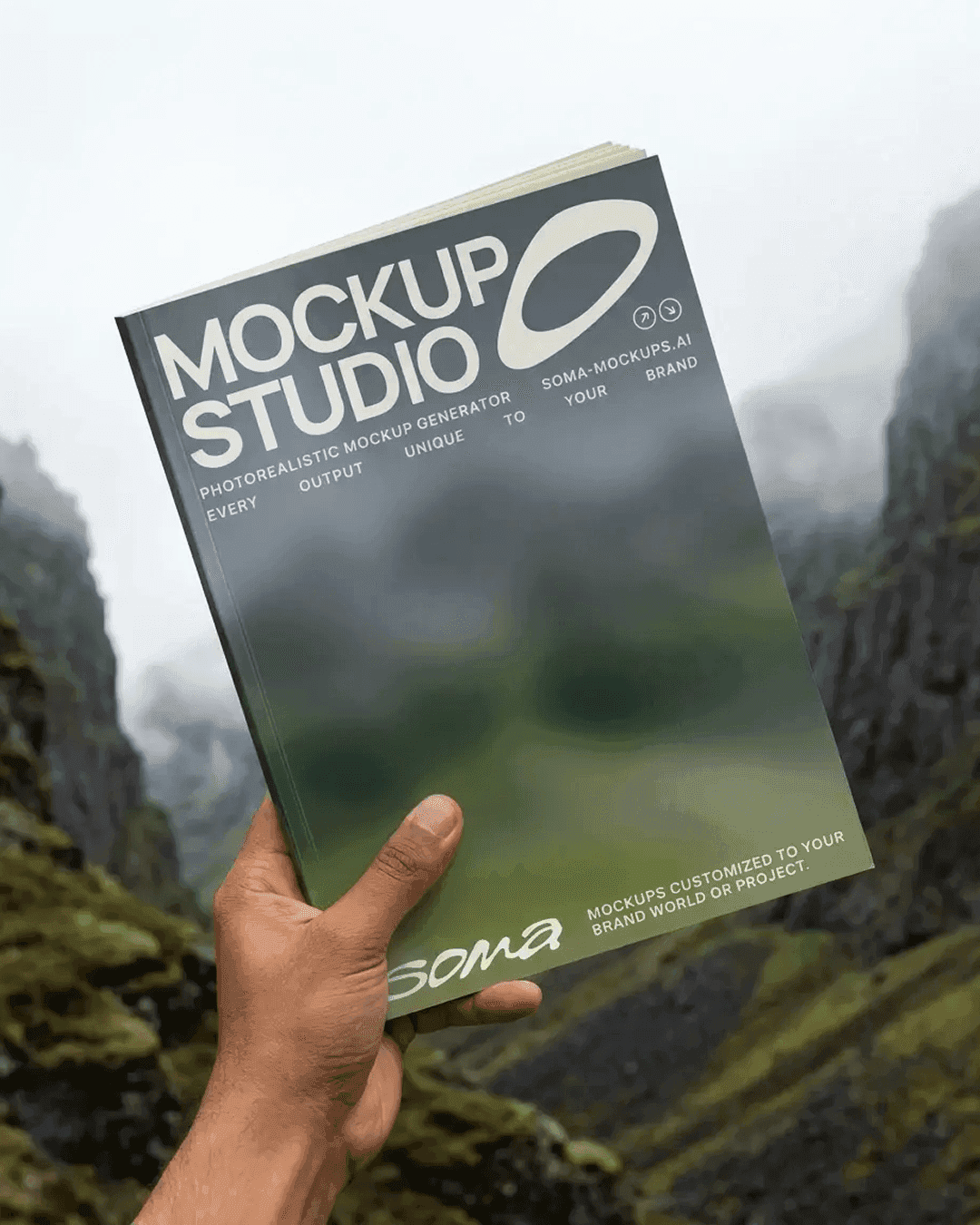 Hand holding a printed Soma "Mockup Studio" magazine in front of a misty mountain landscape