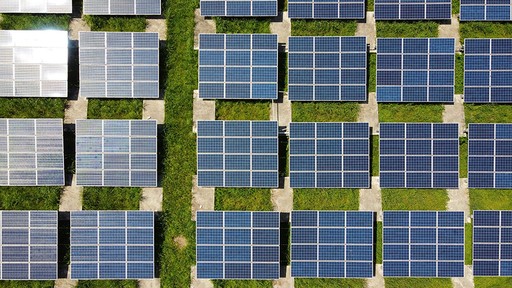An aerial view shows a grid of blue-grey solar panels neatly arranged on vibrant green grass, reflecting sunlight.