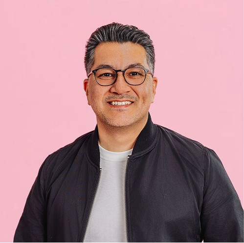 Smiling man wearing glasses and a dark jacket, standing against a soft pink background.