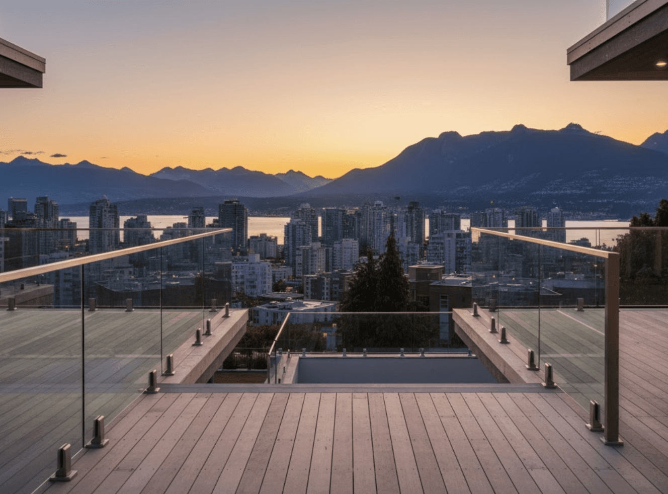 A modern Vancouver waterfront home featuring a sleek glass railing system overlooking the city skyline and mountains at sunset. The scene captures two styles side by side—frameless glass railings with minimal hardware on one deck, and semi-frameless aluminum-post railings on another. The glass reflects warm evening light, showing a clean, expansive view that makes the deck feel larger. Subtle architectural details like 12mm glass panels, stainless-steel fittings, and light wood flooring highlight precision craftsmanship and luxury construction. The overall mood conveys premium design, safety, and sophistication—ideal for a high-end Vancouver residential renovation feature.