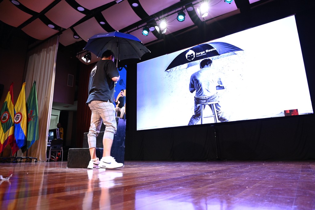 Jorge Baena en el escenario sosteniendo un paraguas negro, frente a una gran pantalla que muestra la misma imagen del paraguas como parte de su presentación.