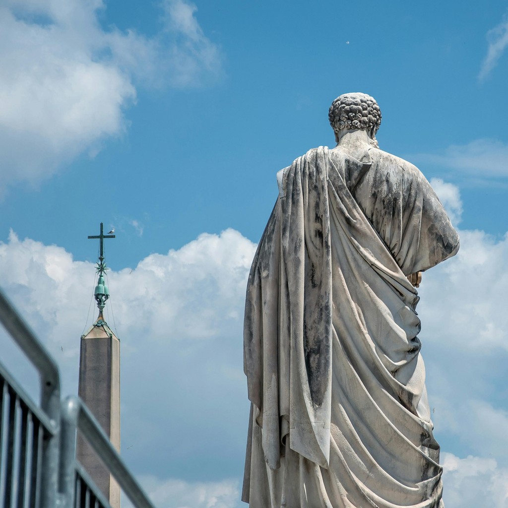 The image depicts the back of a marble statue of St. Peter, draped in traditional Roman attire, gazing towards the obelisk and the cross atop St. Peter's Square in Vatican City. The statue is set against a backdrop of a vibrant blue sky with scattered clouds, symbolizing the religious and historical significance of the location.