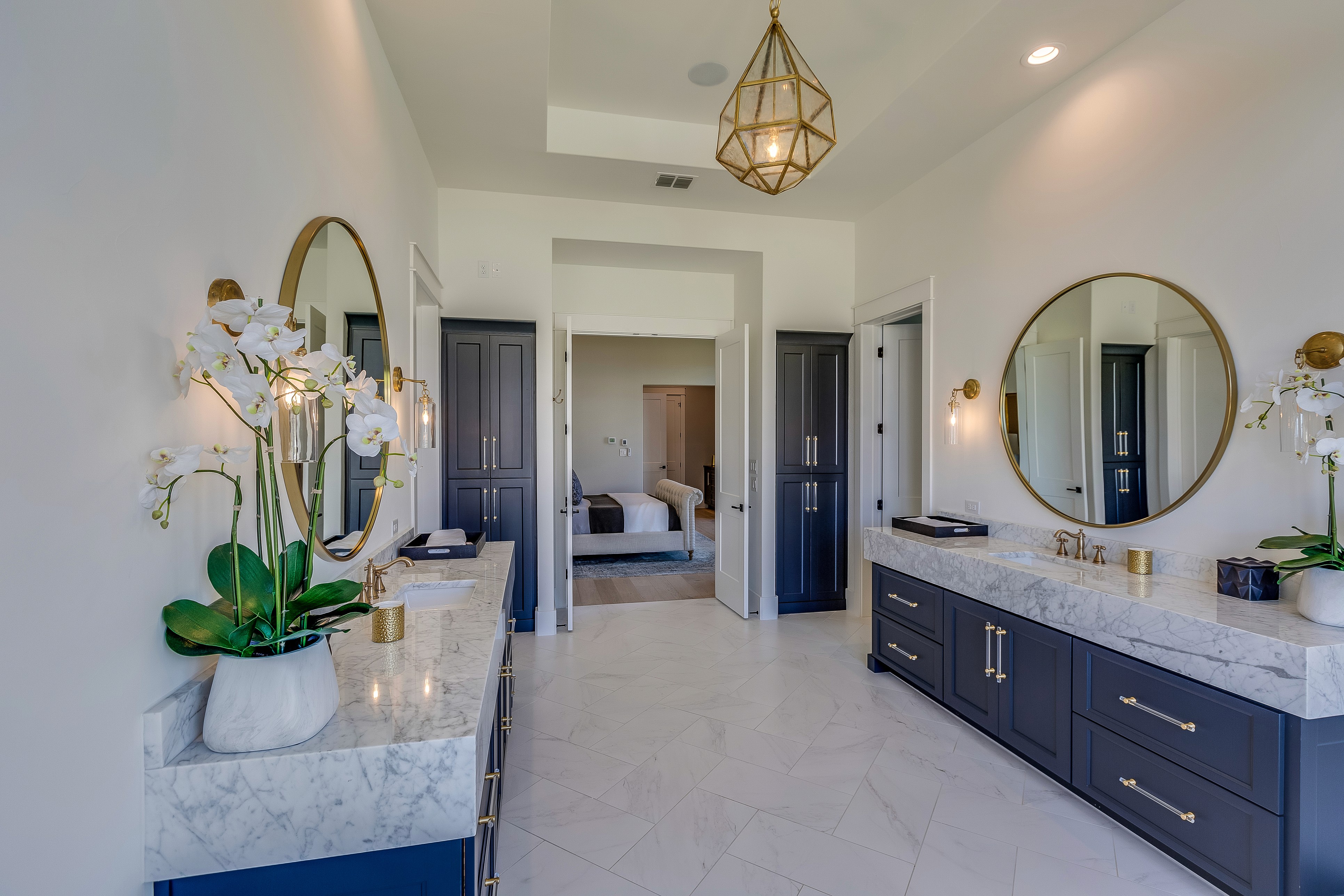 Large primary bathroom with blue double vanitites, thich marble countertops, gold accent fixtures, and herringbone pattern floor tile