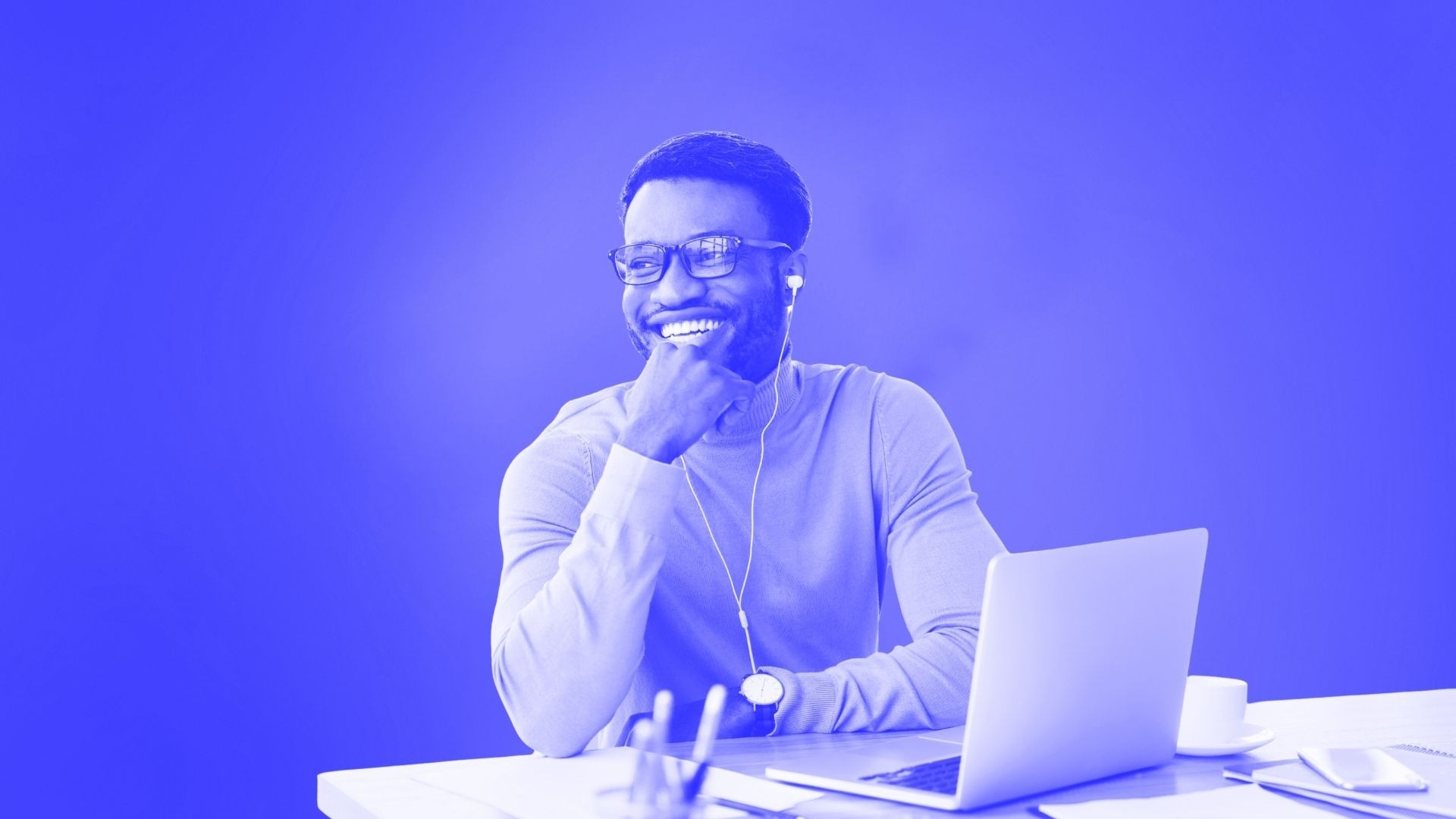 A smiling man sitting down in front of a laptop at a table wearing headphones