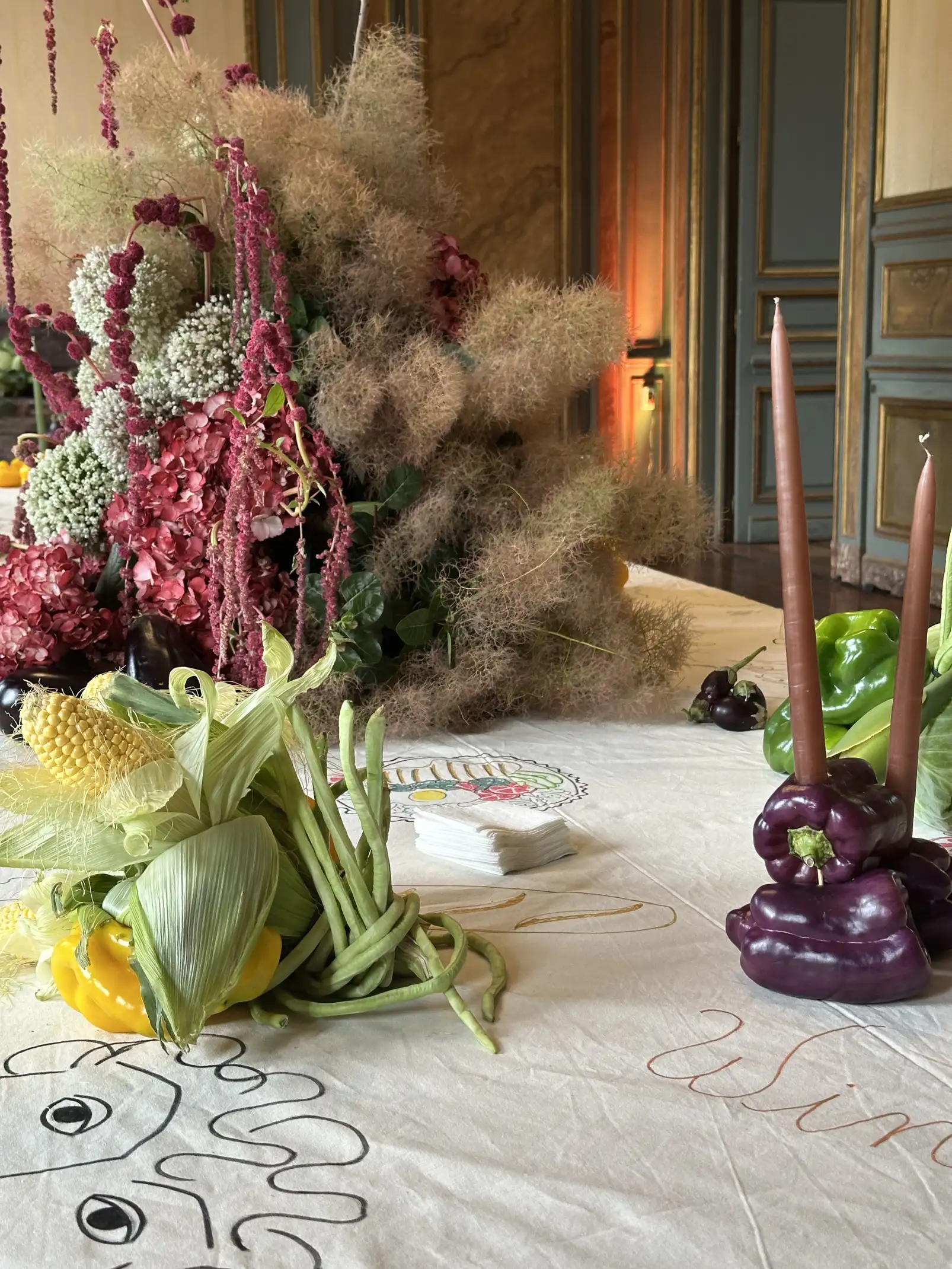 Centro de mesa floral con hortensias rosadas, flores blancas y amaranto rojo, acompañado de una composición de vegetales frescos como maíz, pimientos y chauchas, sobre un mantel blanco ilustrado, en un salón de estilo clásico con paredes decoradas
