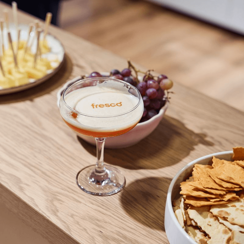 Cocktail in a coupe glass with foam branded "fresco" sits on a wooden table beside grapes, chips, and snacks