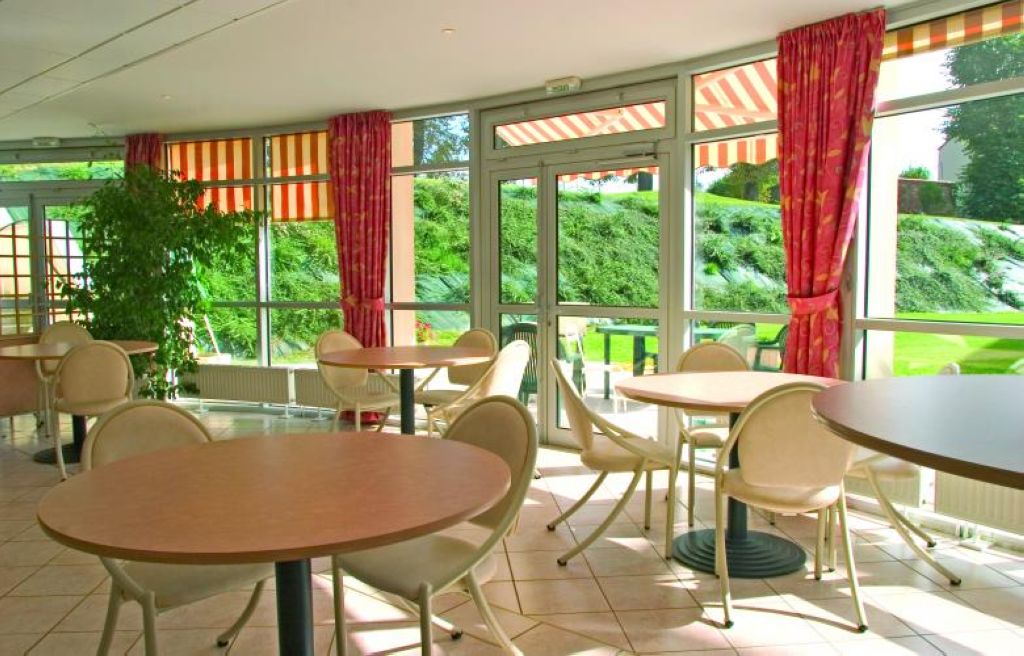 Interior common area with wooden furniture, green plants, red support columns, and bright natural light coming through the windows
