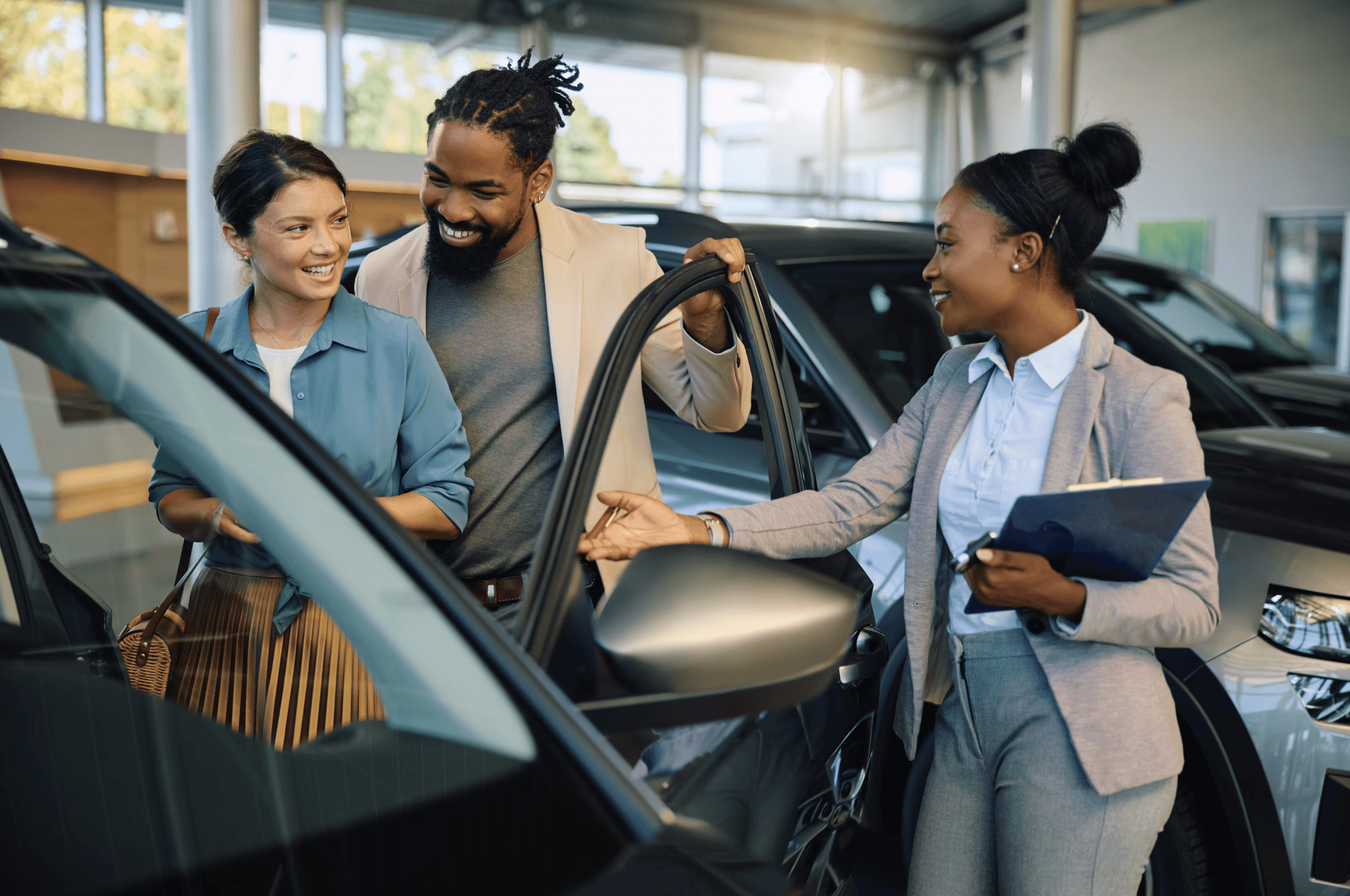 Smiling couple exploring a new car at a dealership with assistance from a friendly saleswoman holding a clipboard.