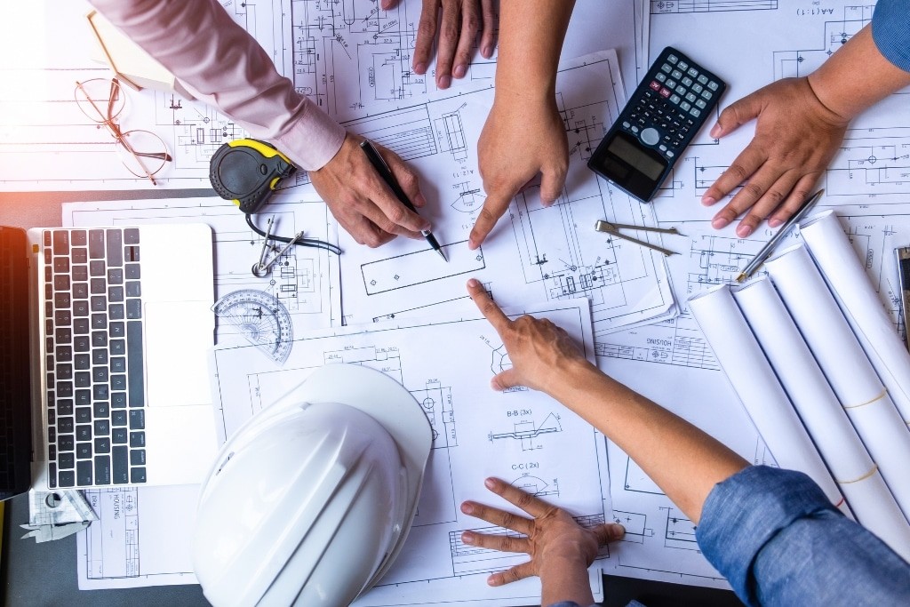 Three construction workers in safety vests and helmets review blueprints at a building site. The setting is sunlit, with a partially-built structure in the background.