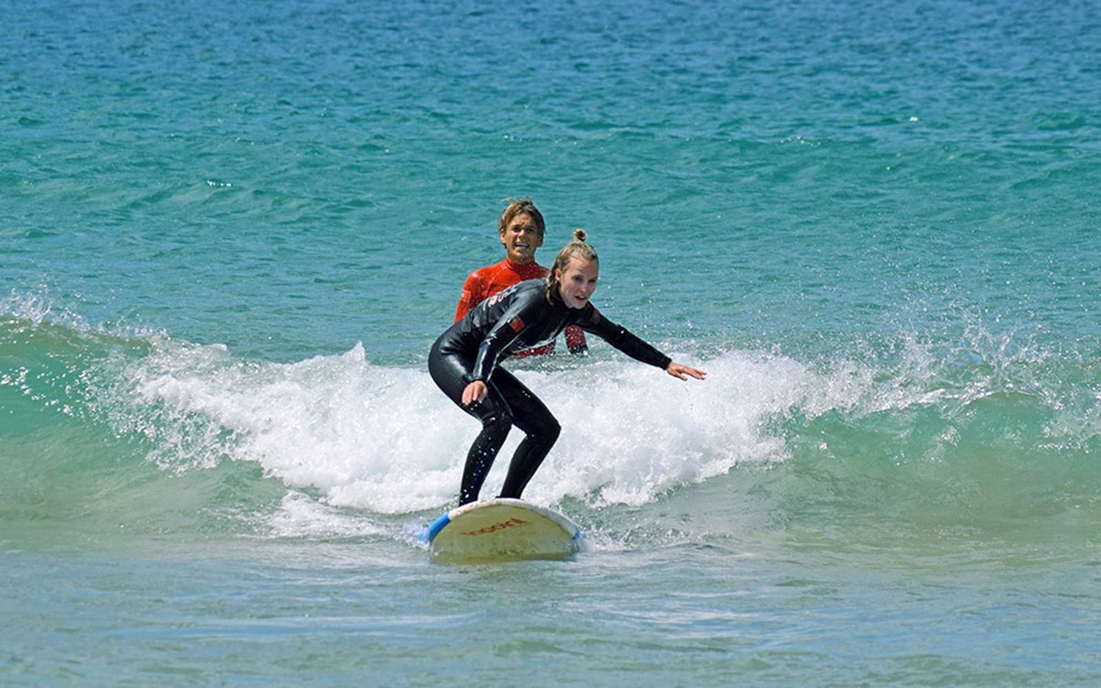 Öğretmen eşliğinde, bir öğrencinin dalgada yönlendirilmesiyle gerçekleştirilen grup sörf dersi.