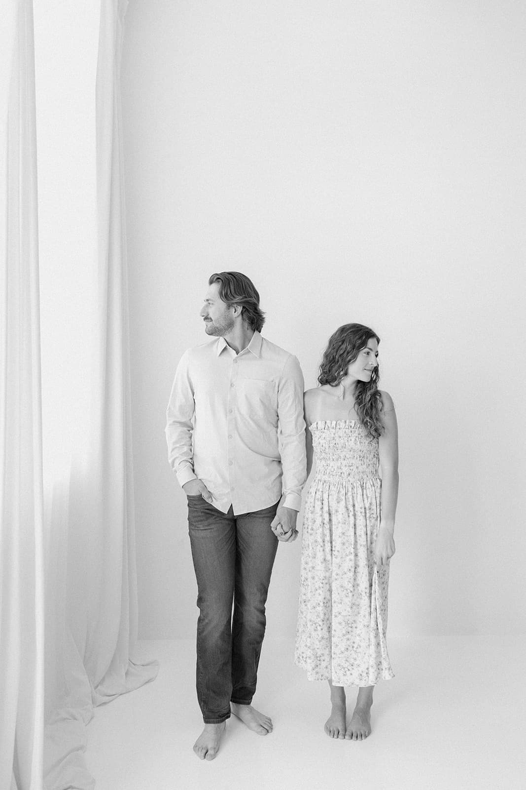 A couple standing in front of a large window at Revelator Studio in Shreveport holding hands, looking in opposite directions.