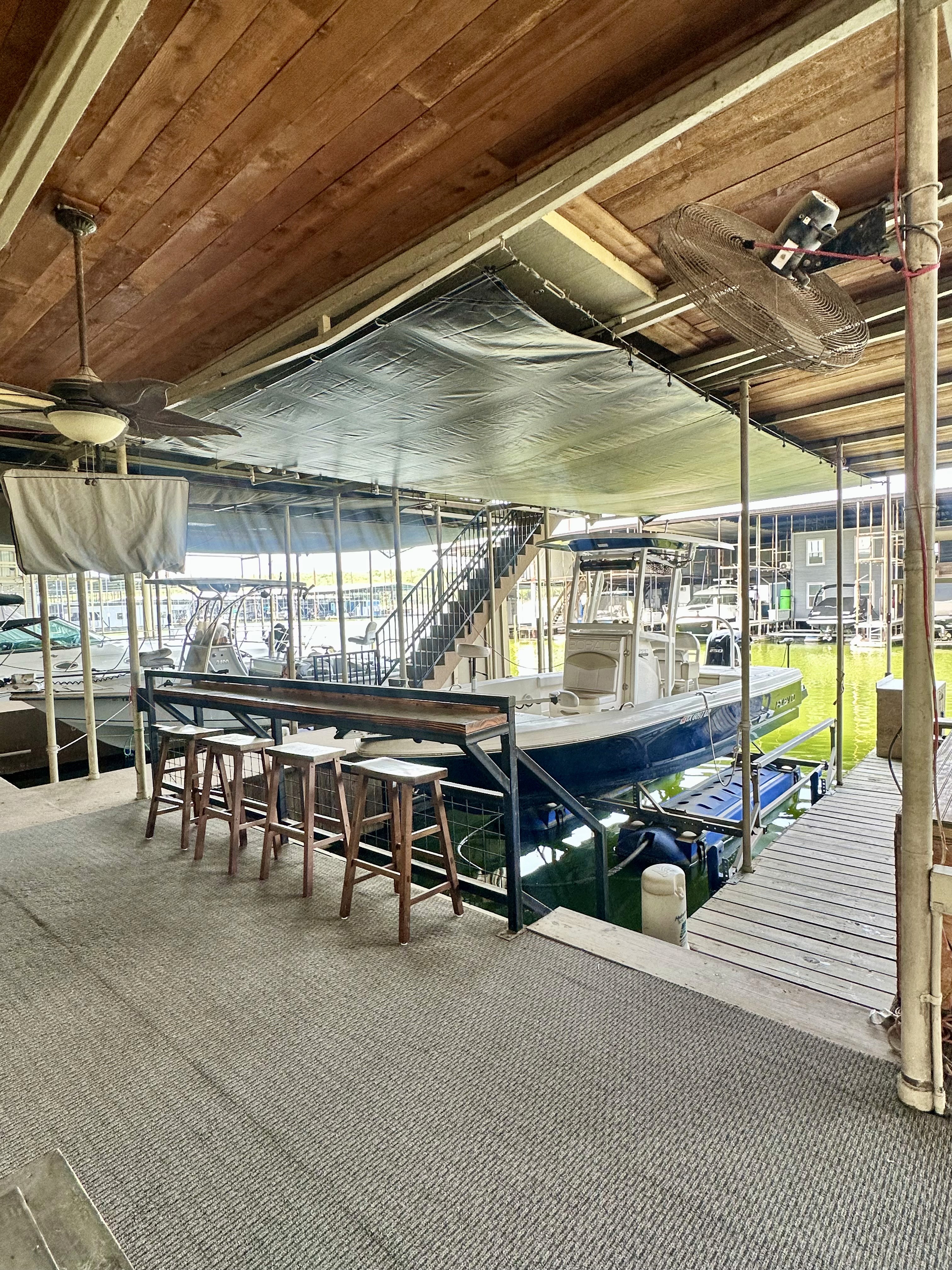 A covered boat dock features a white motorboat moored beside a wooden bar with stools, surrounded by various other docked boats, and sheltered by a slanted wooden ceiling and fan.