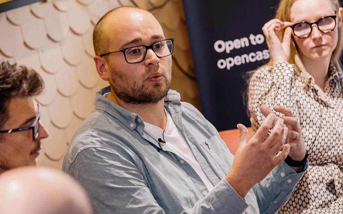 A panelist speaks while gesturing, seated alongside others in a moderated discussion. Event branding and soft lighting frame the conversation.
