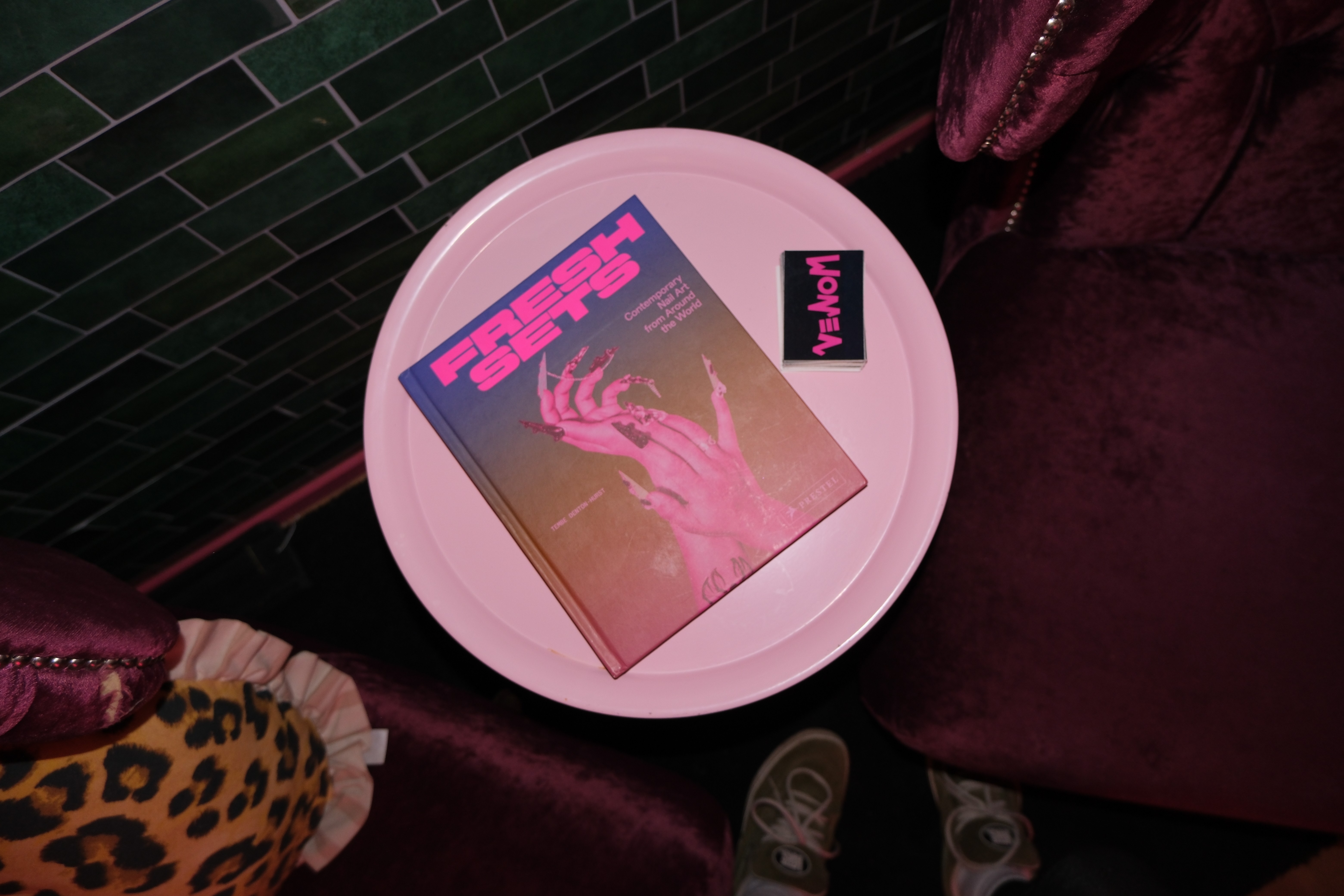 A top-down shot of a white round side table holding a book titled Fresh Sets: Contemporary Nail Art from Around the World and a small black card with the pink "VENOM" logo.