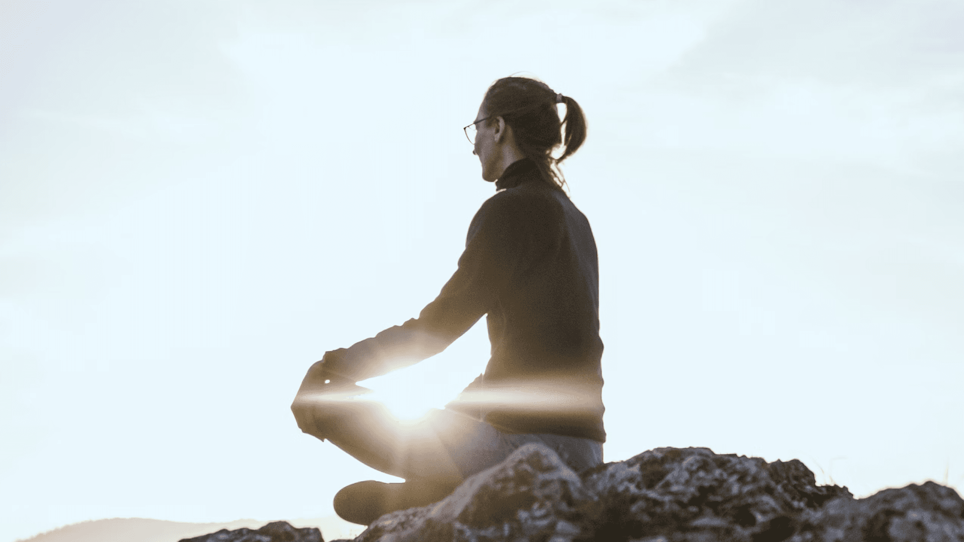 A woman meditating on a rocky hill at sunrise, symbolizing mindfulness.