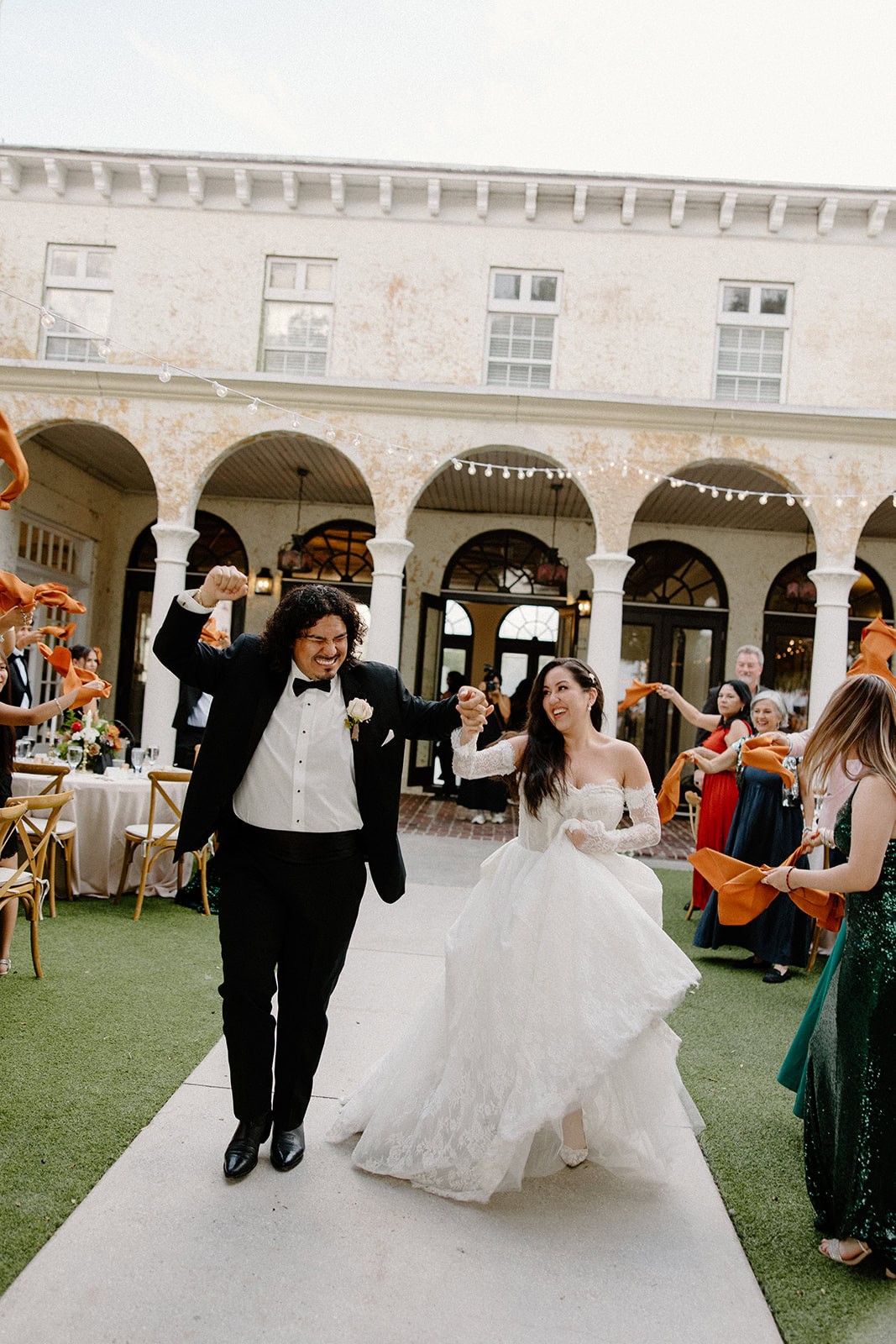 A man in a tuxedo and a woman in a white dress are walking forward, smiling and looking at each other. They are surrounded by their wedding party on a grassy lawn.