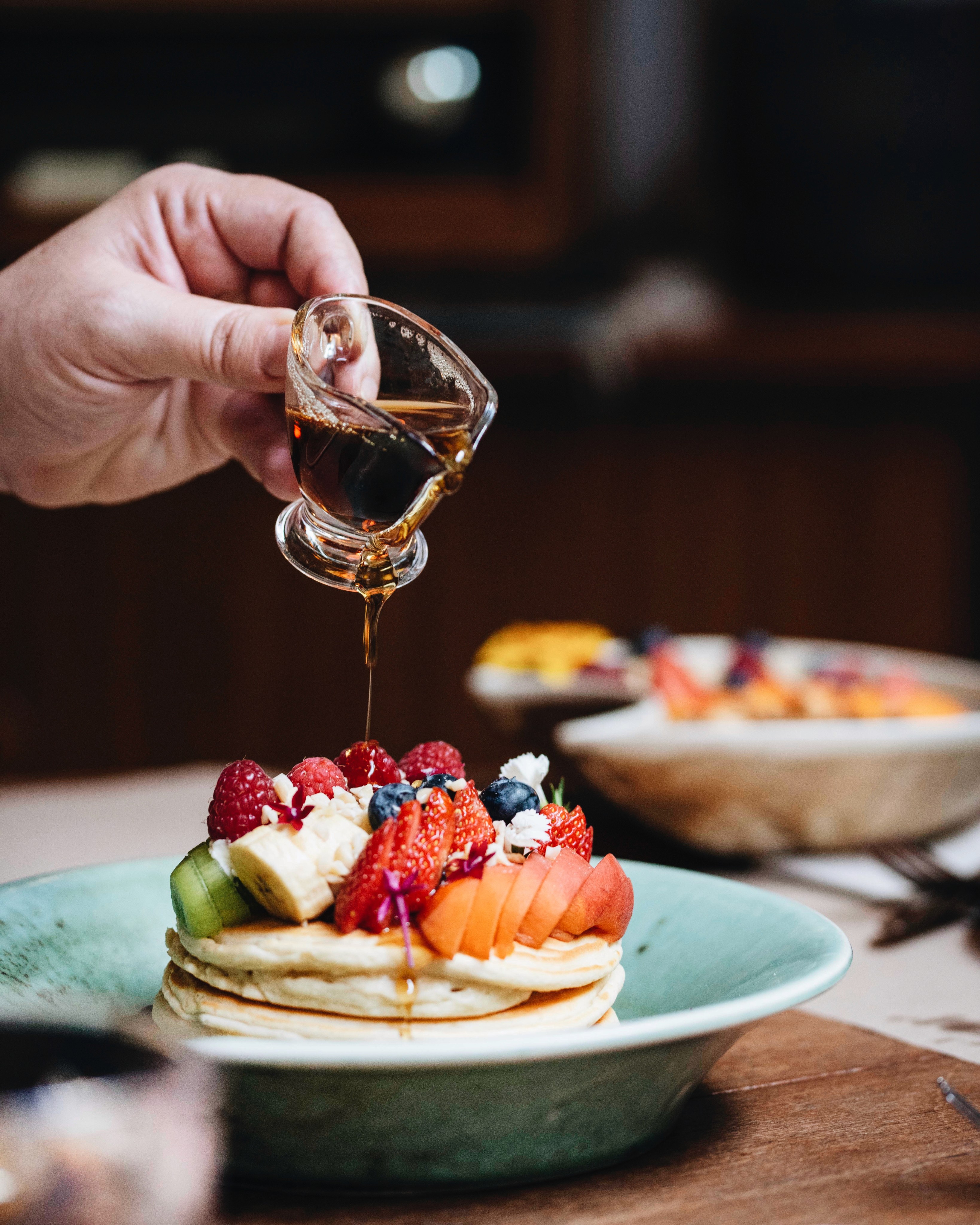 Pouring maple syrup over fluffy pancakes topped with fresh raspberries, strawberries, kiwi and edible flowers — vegetarian brunch at Faire. Brunch & Drinks, Eixample Barcelona