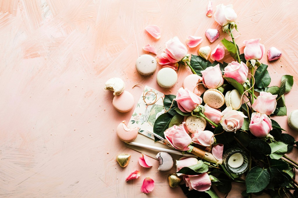 A romantic flat lay featuring soft pink roses, assorted pastel macarons, and heart-shaped chocolates, arranged on a pink textured background. Perfect for Valentine’s Day, anniversaries, or romantic celebrations.