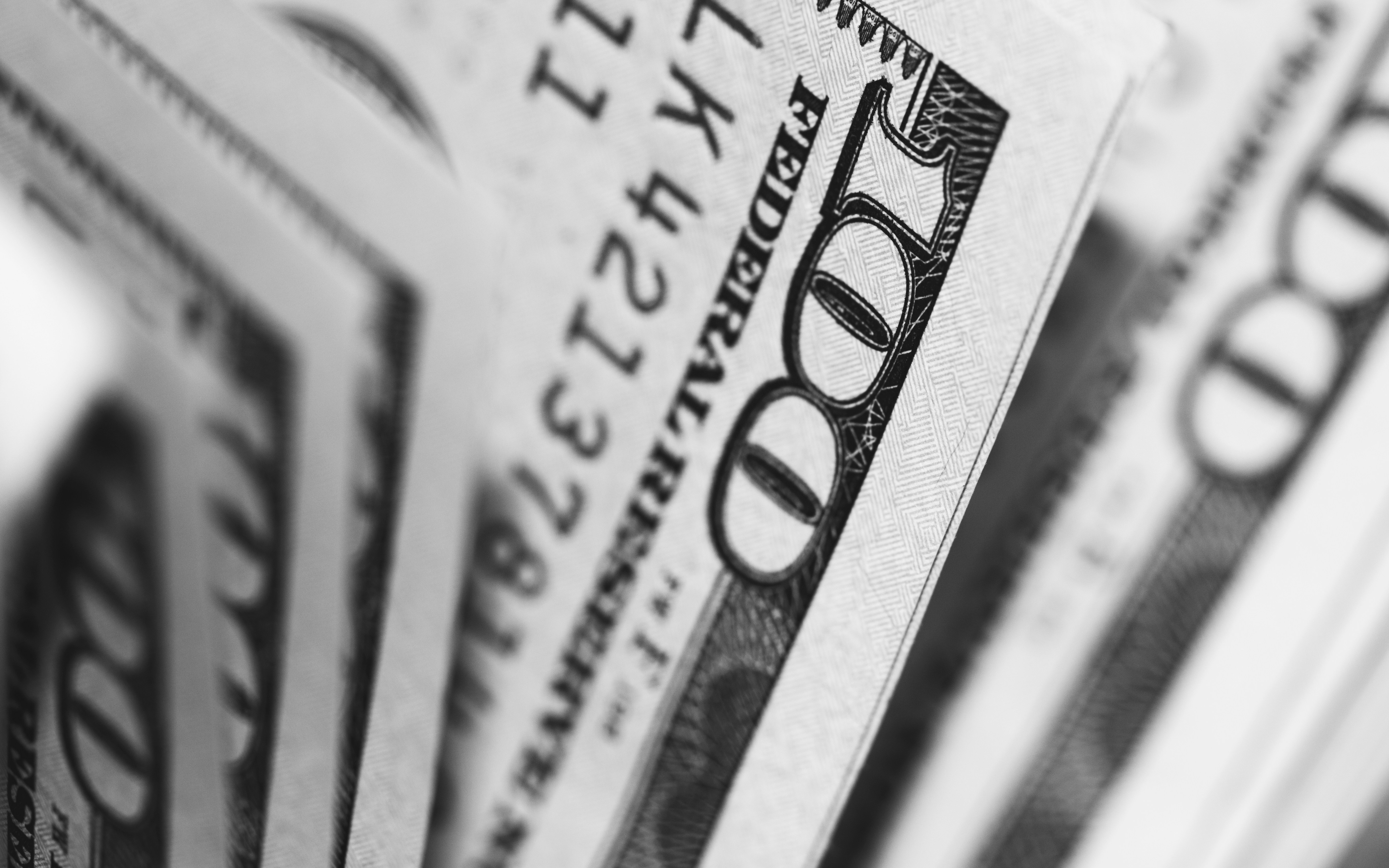 A close-up black and white photograph of several one-hundred-dollar bills, fanned out and partially overlapping, with the focus on the "100" in the foreground. The image emphasizes the concept of money and finance.