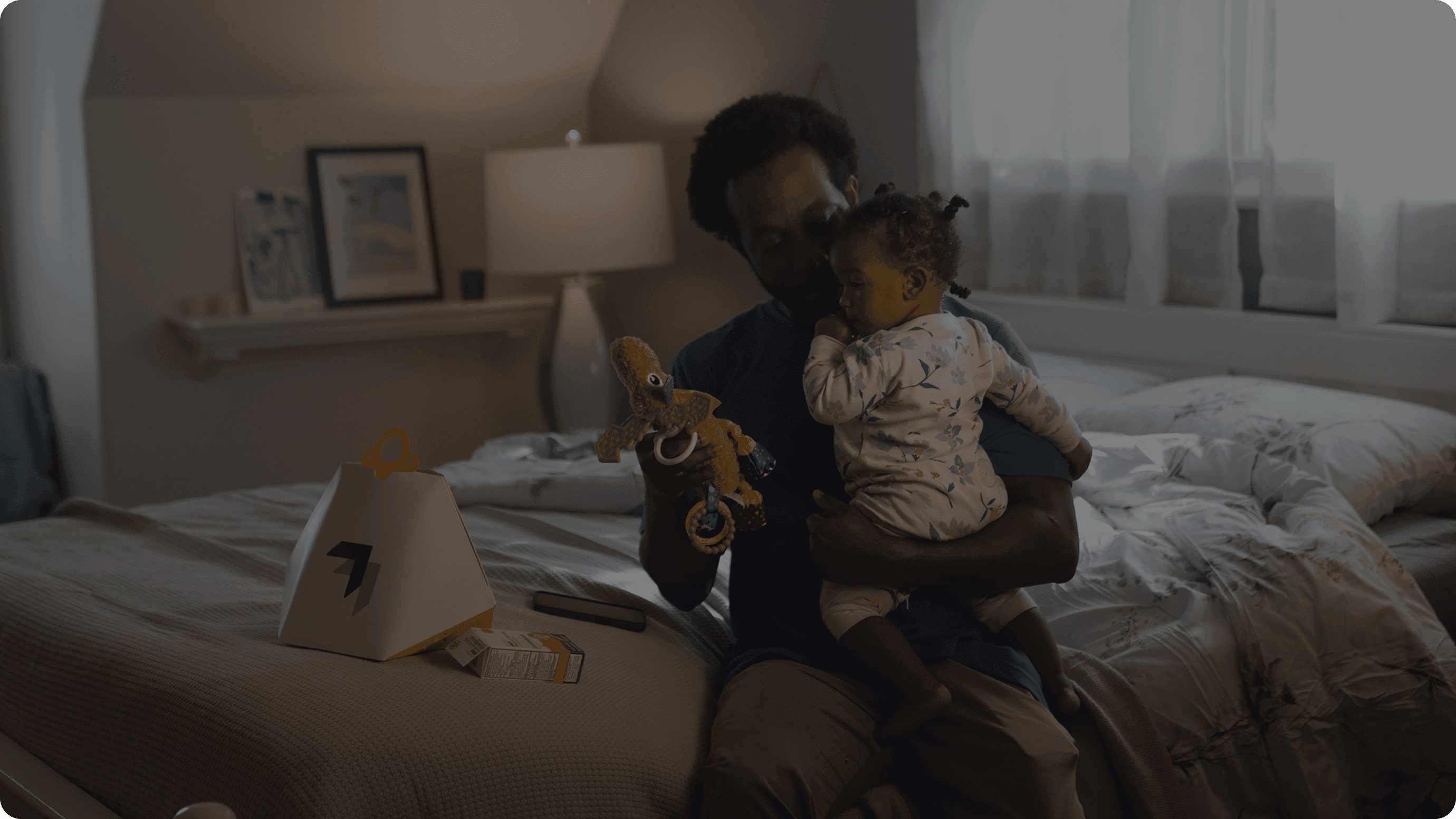 A person is sitting on a bed in a dimly lit bedroom, holding a small child and a plush toy, with a fast-food takeout bag visible on the bed.