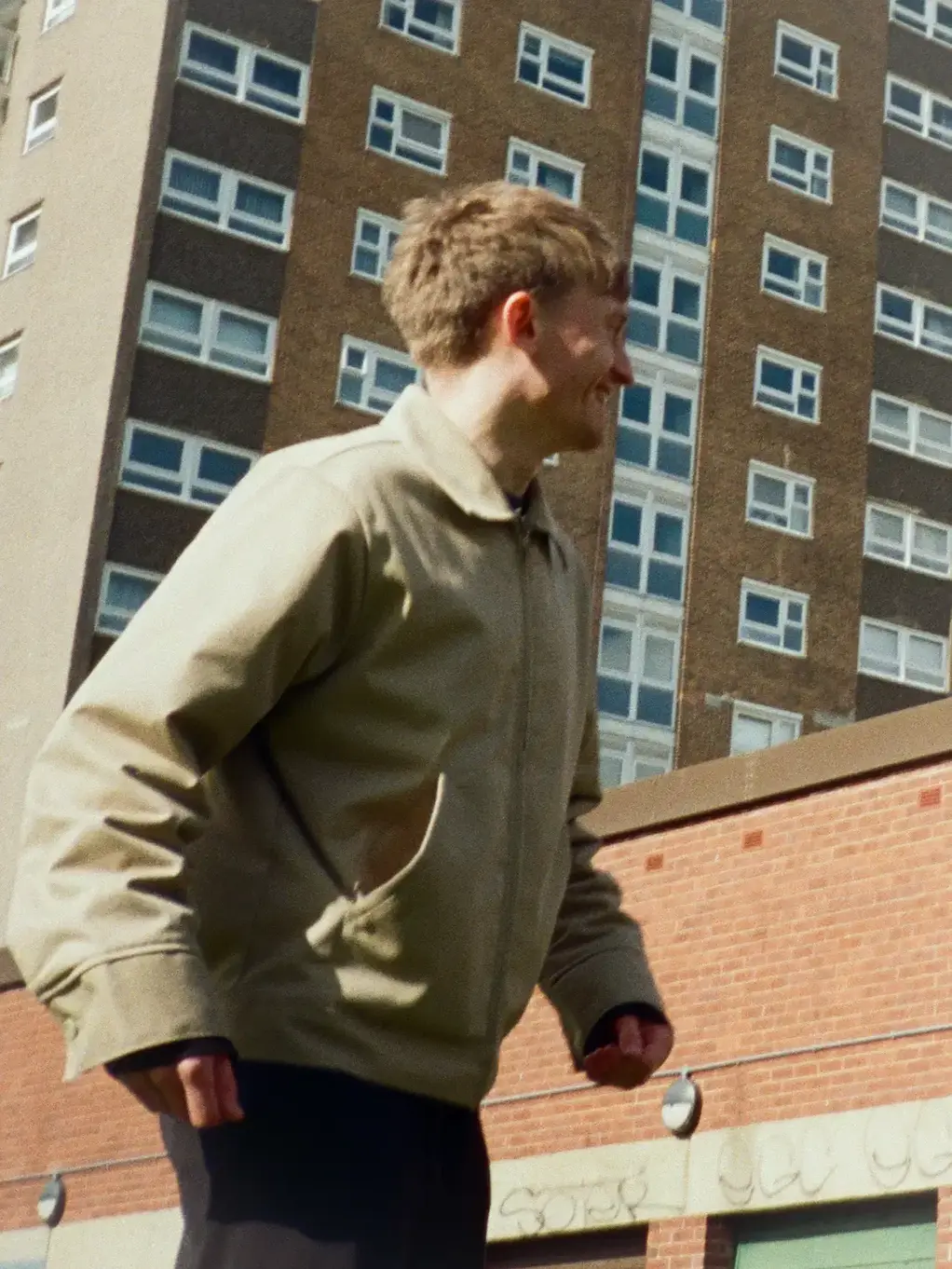 A person wearing a beige jacket is walking in front of a tall, brown brick apartment building with numerous windows, under a clear sky.