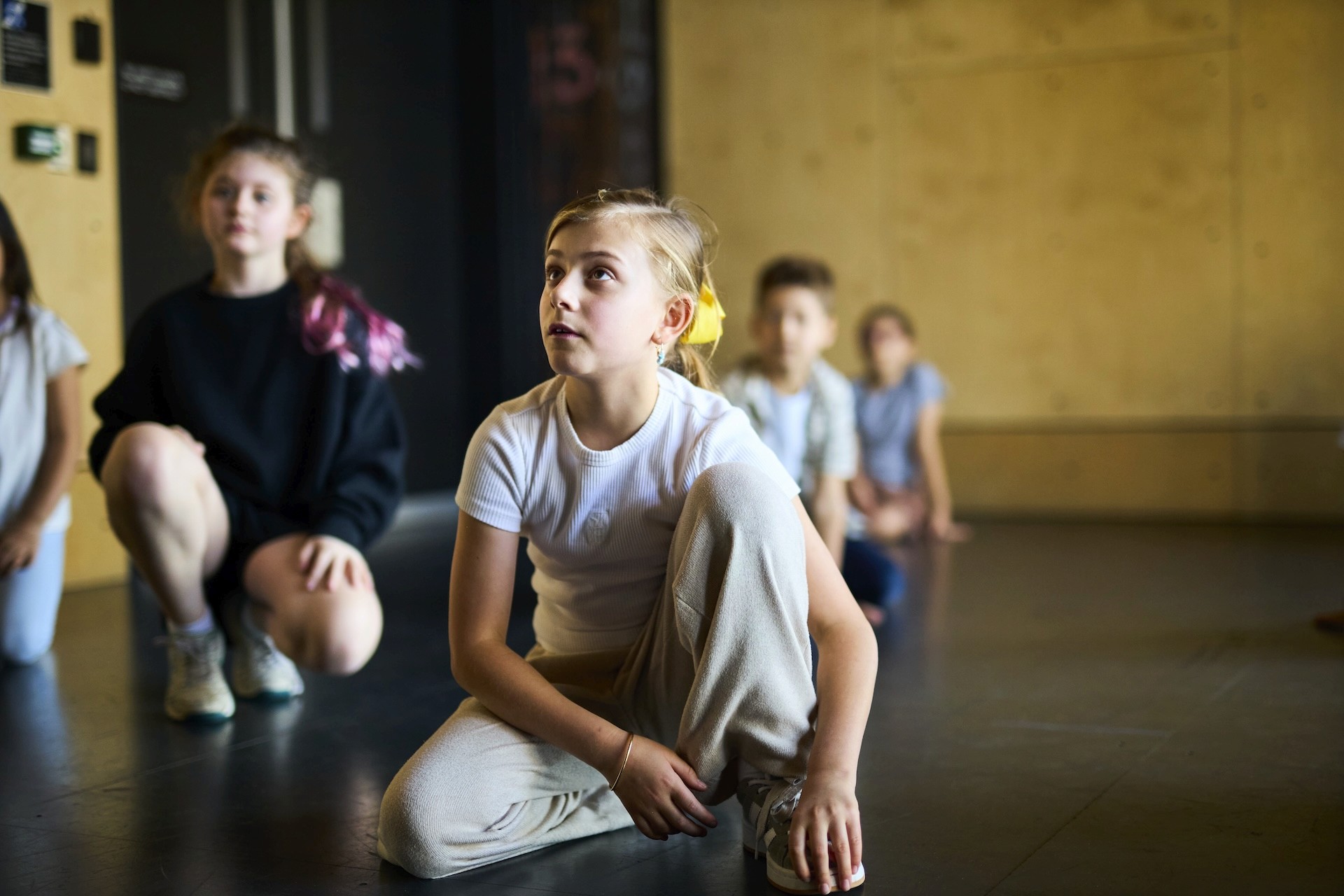 Children sit and kneel on the floor of a studio space, listening intently during a drama workshop.