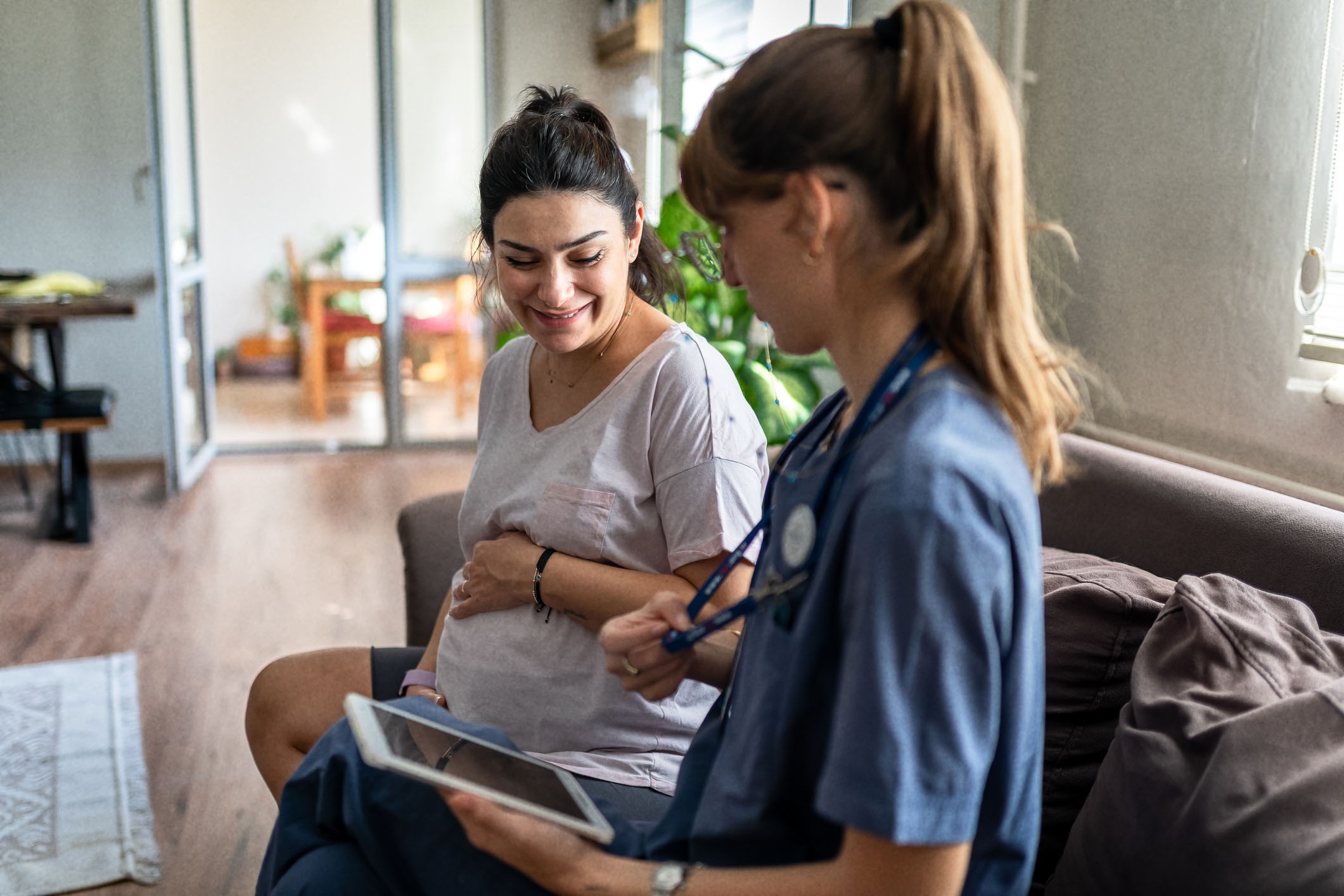 a pregnant woman is sitting next to a haealthcare provider on a sofa. The provider is looking at a tablet computer