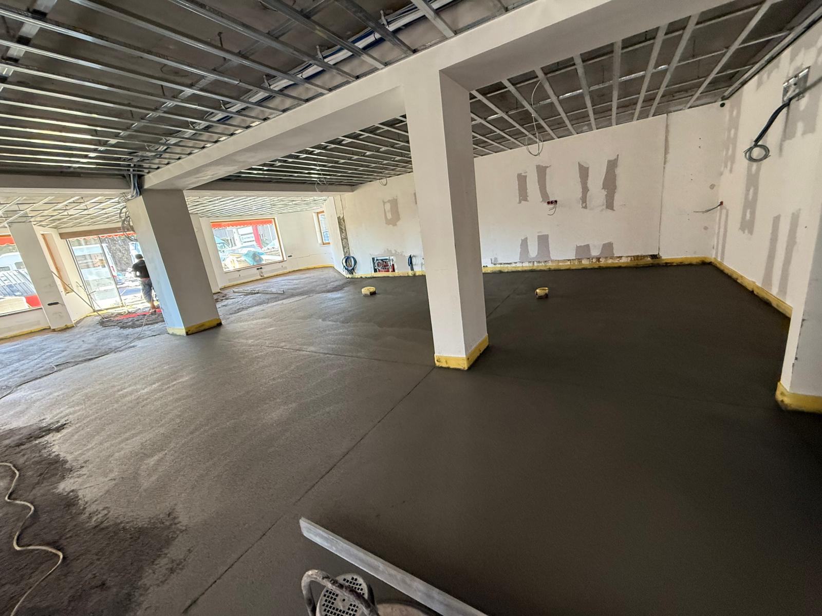 Innenansicht einer Baustelle mit frischem Betonboden, freiliegender Deckenkonstruktion und Stützpfeilern.
