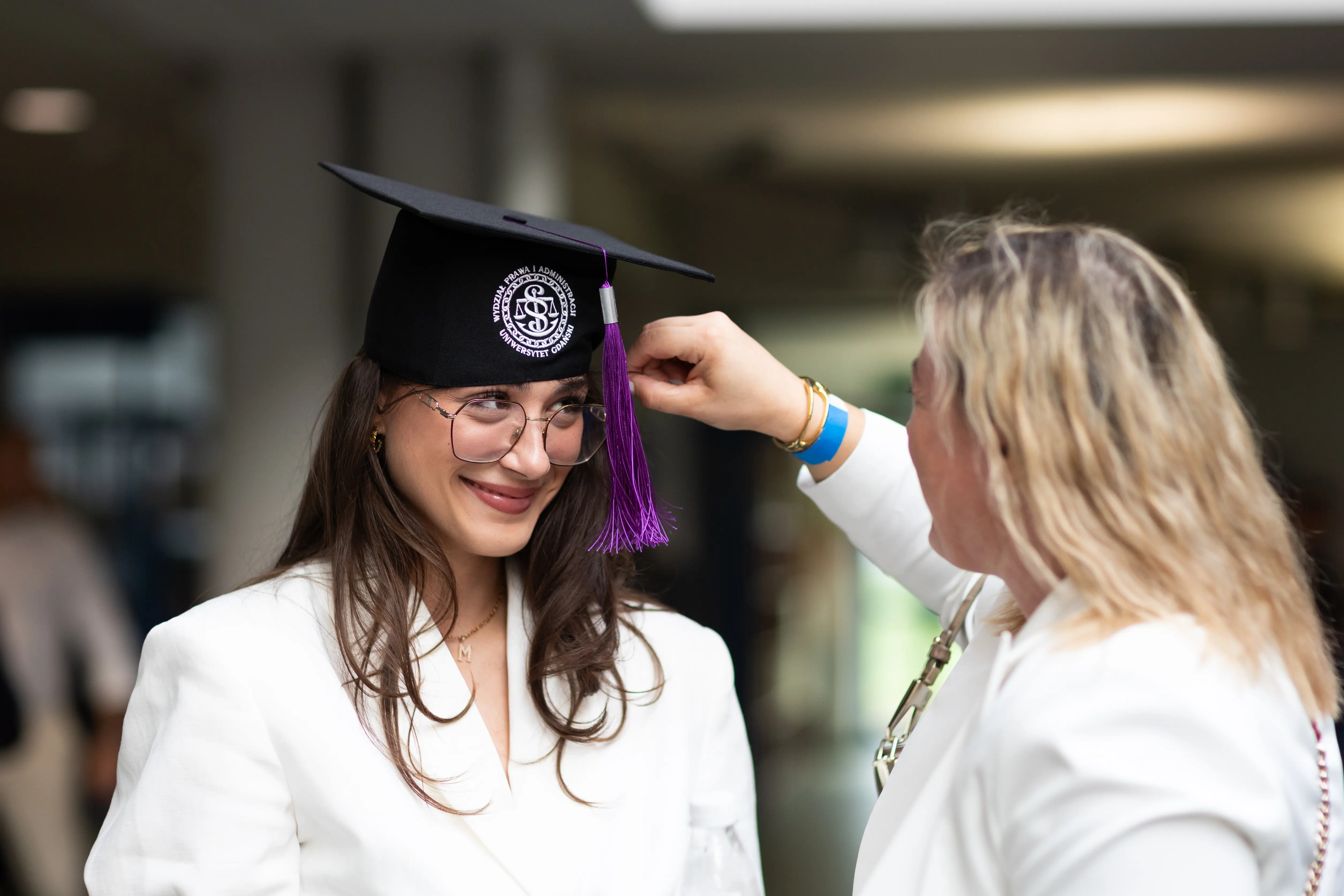 Młoda kobieta z uśmiechem bierze udział w ceremonii wręczenia dyplomów, gdy ktoś poprawia jej czapkę absolwenta. Obie osoby ubrany są w białe stroje.
