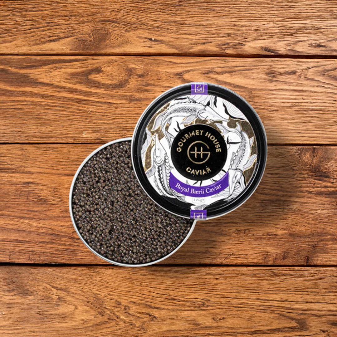 Top-down view of an open 125g tin of Gourmet House Royal Baerii Caviar. The tin is filled with small, dark sturgeon eggs and sits on a horizontal wooden plank background. The lid, featuring sturgeon illustrations and a purple label, rests partially over the tin.