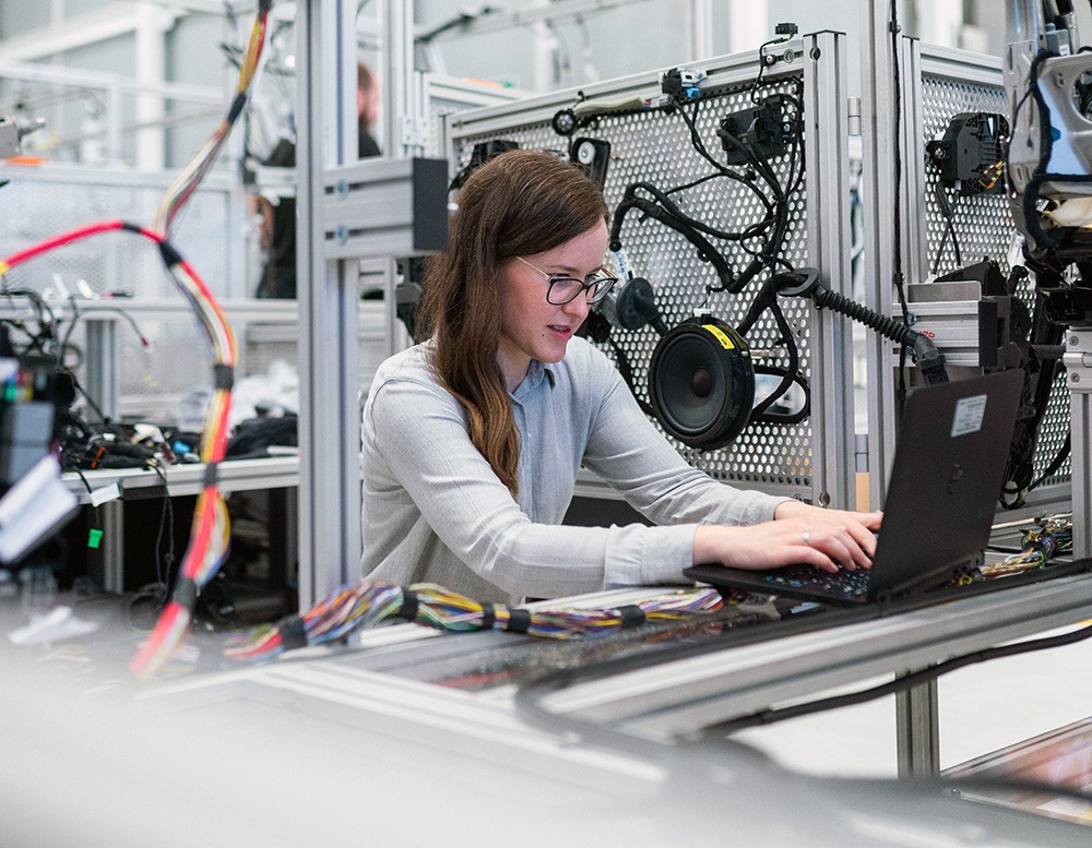 A woman wearing glasses works at a laptop in a technical laboratory or manufacturing facility. She sits at a workbench surrounded by electronic testing equipment, cables, and industrial machinery, representing women in STEM fields and technical careers.