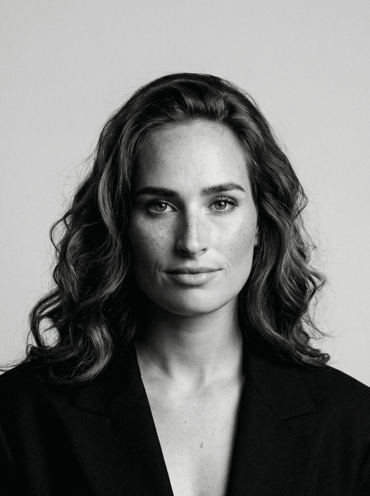 Man with short hair and beard wearing a dark blazer and black shirt against a plain background.