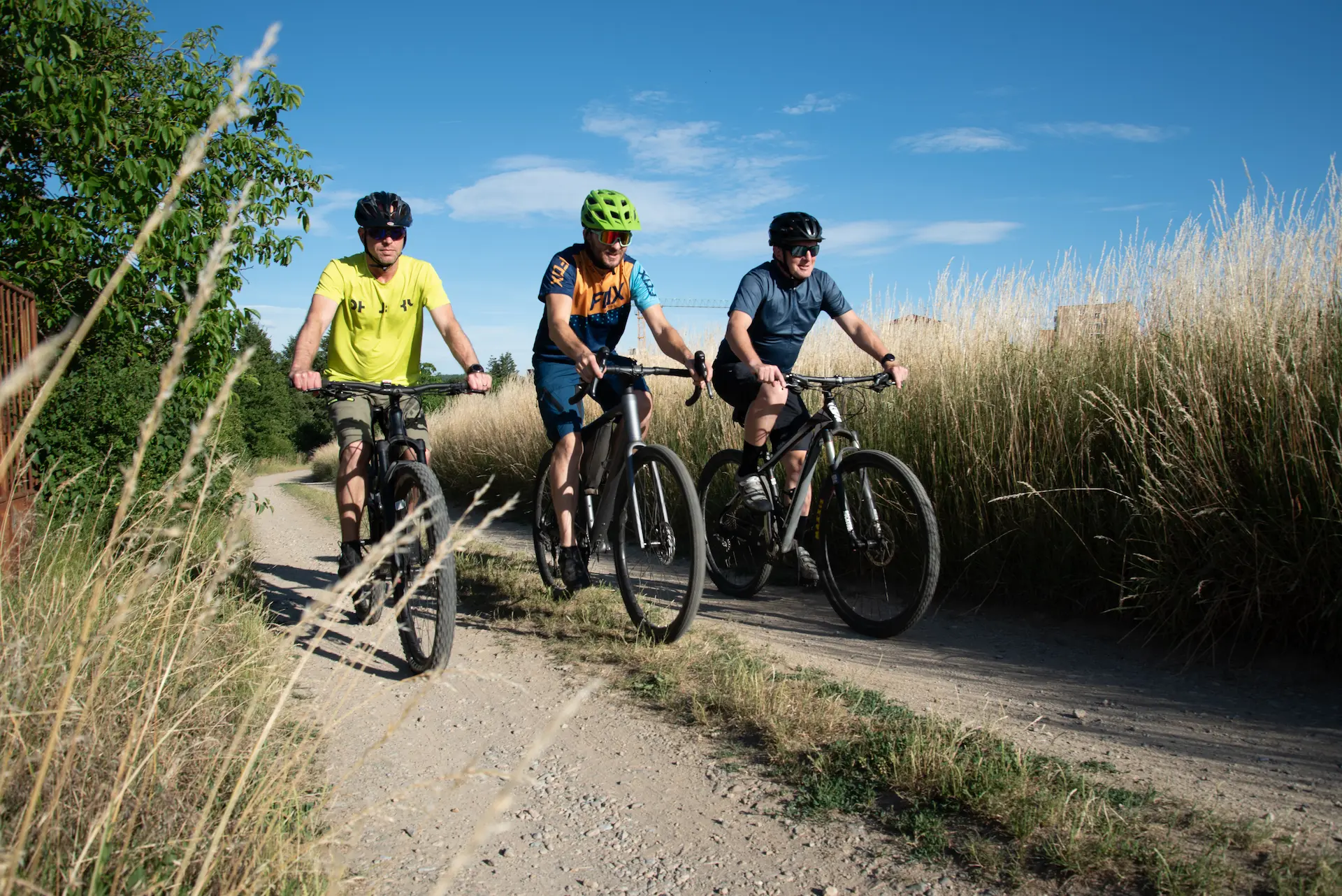 Tři cyklisté na horských kolech jedou po štěrkové cestě mezi vysokou trávou