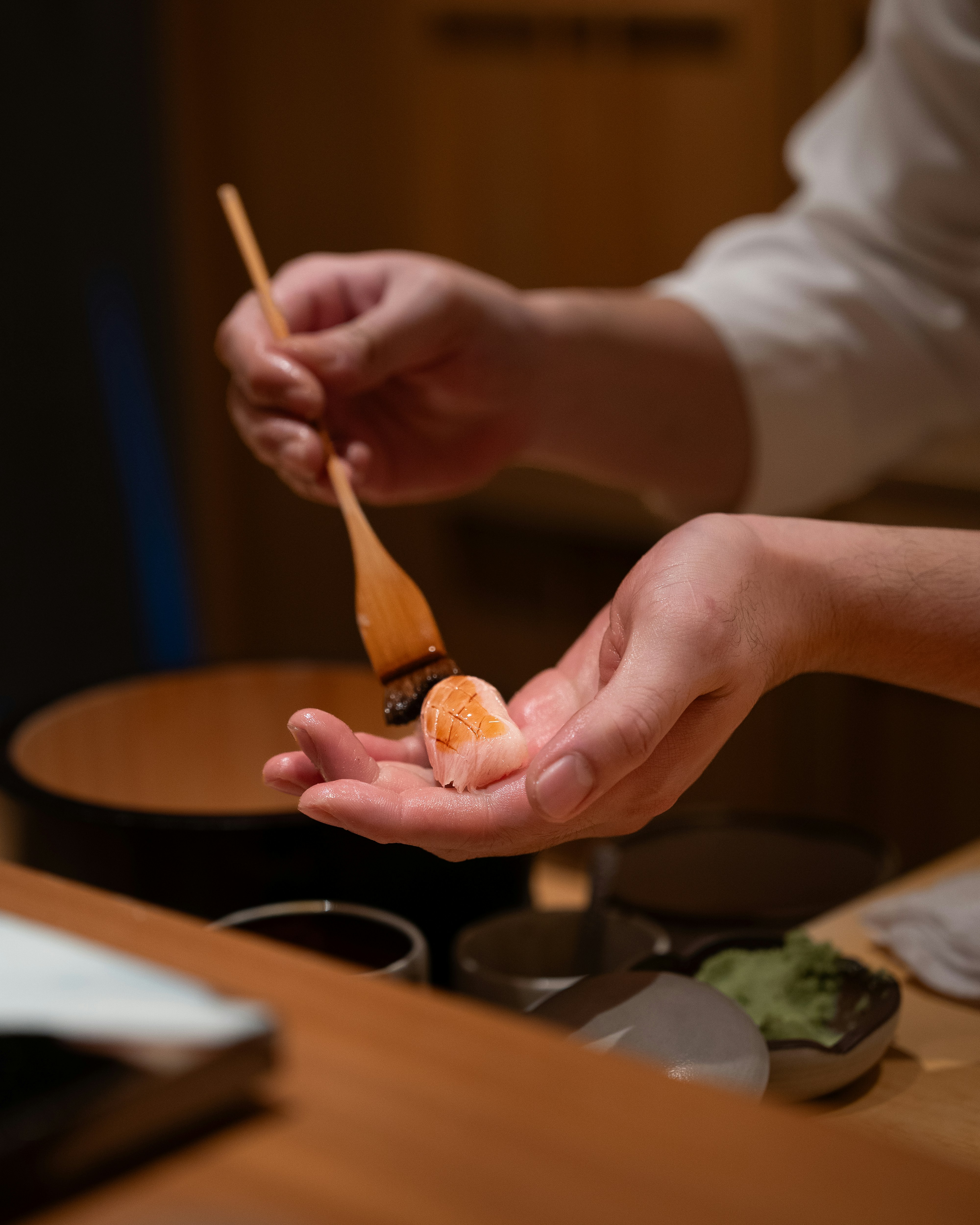 A sushi chef brushing soy onto a piece of nigiri held in his palm at a wooden counter