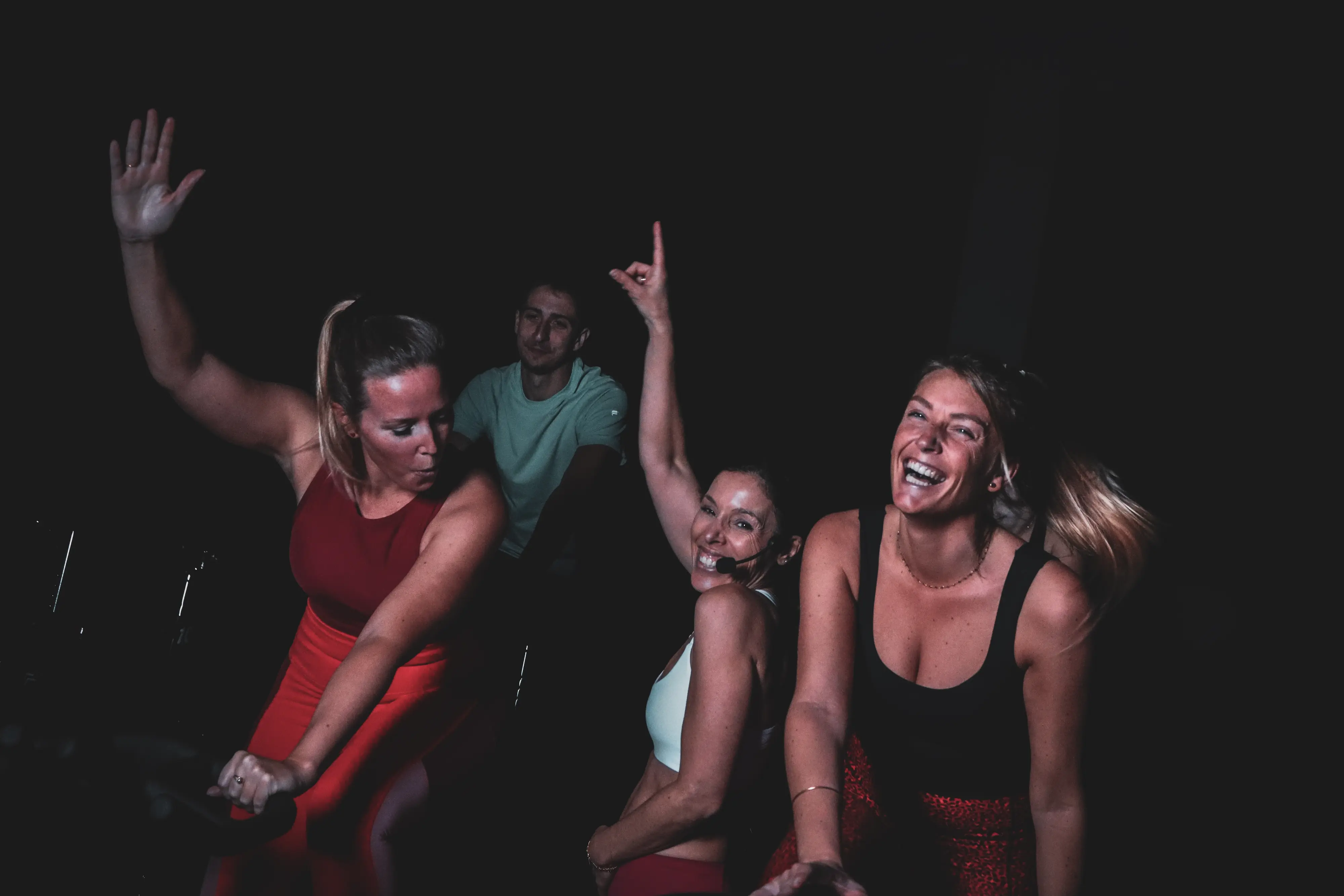 Trois participantes et une coach en pleine séance de cycling, sourires et bras levés dans la pénombre du studio