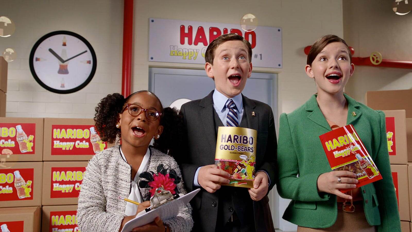 Three children dressed as adults holding HARIBO products stand in front of boxes and a clock, appearing to sing or speak enthusiastically in a brightly lit room.