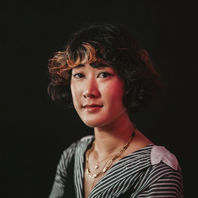 Photo portrait de profil de la designer et directrice artistique Diana SOM. Une jeune femme aux cheveux noirs bouclés courts avec des reflets cuivrés, portant une chemise rayée noire et blanche et des colliers dorés. Le portrait est réalisé sur un fond noir avec un éclairage studio doux et chaleureux.