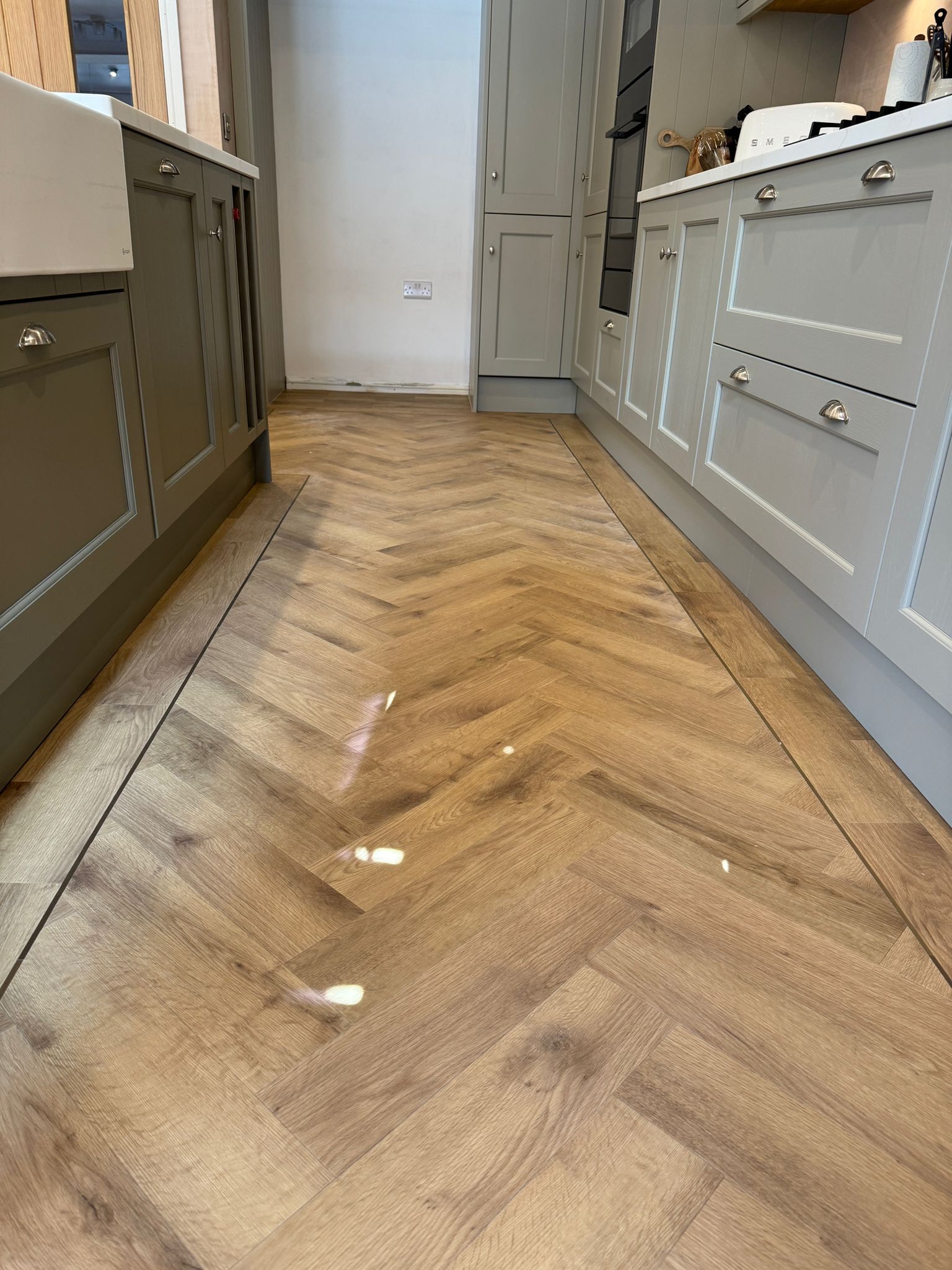 A modern kitchen with light gray cabinets features a stunning herringbone pattern laminate vinyl tile (LVT) flooring, showcasing a warm, wood-like finish popular in Sheffield homes.