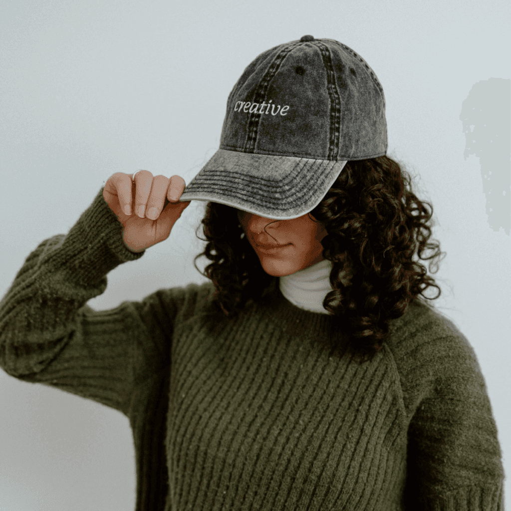 woman with curly hair wearing a black baseball cap that reads "creative", a green sweater, and a white turtleneck