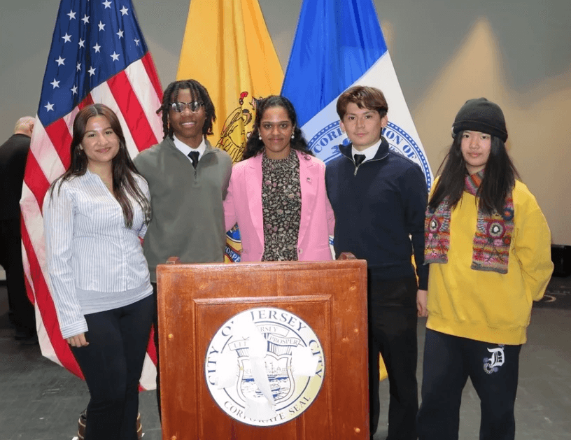 A speaker on stage, illuminated against a dark background, gestures while addressing an audience.