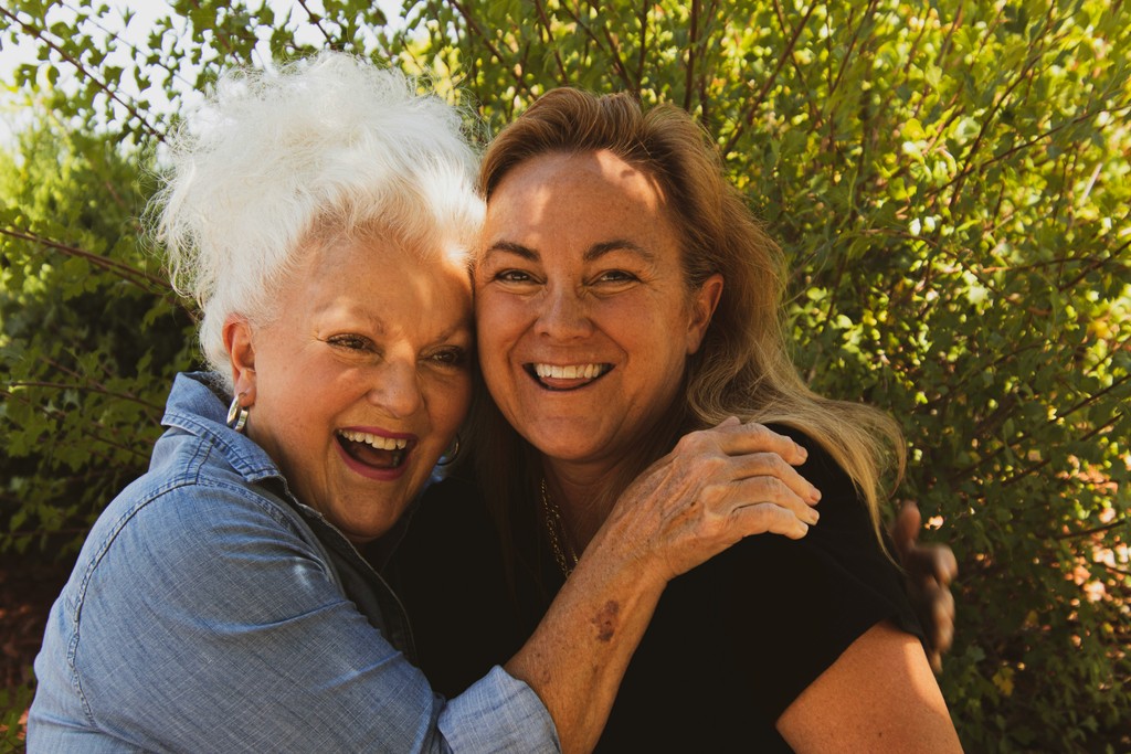 Two joyful women share a warm embrace and laugh together outdoors, showcasing their close bond and happiness in a sunlit, natural setting.