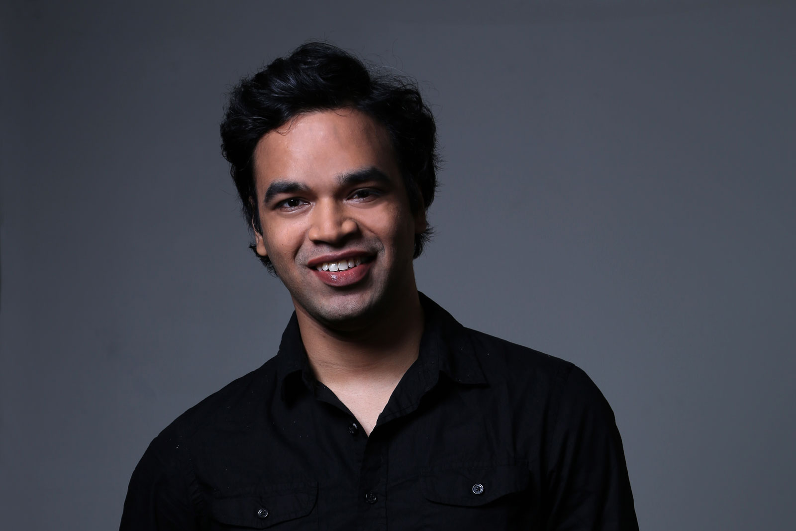 The image shows a man with dark, curly hair and brown skin, wearing a black button-down shirt. He is looking directly at the camera with a slight smile. The background is a solid dark gray color.
