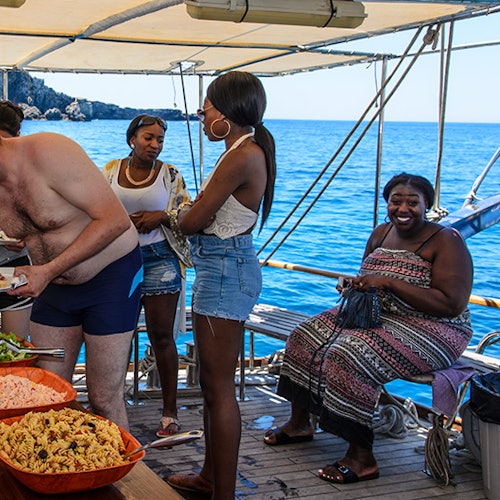 Four people are on a boat with a food spread, smiling and talking under a canopy. The sea and distant rocks are visible.
