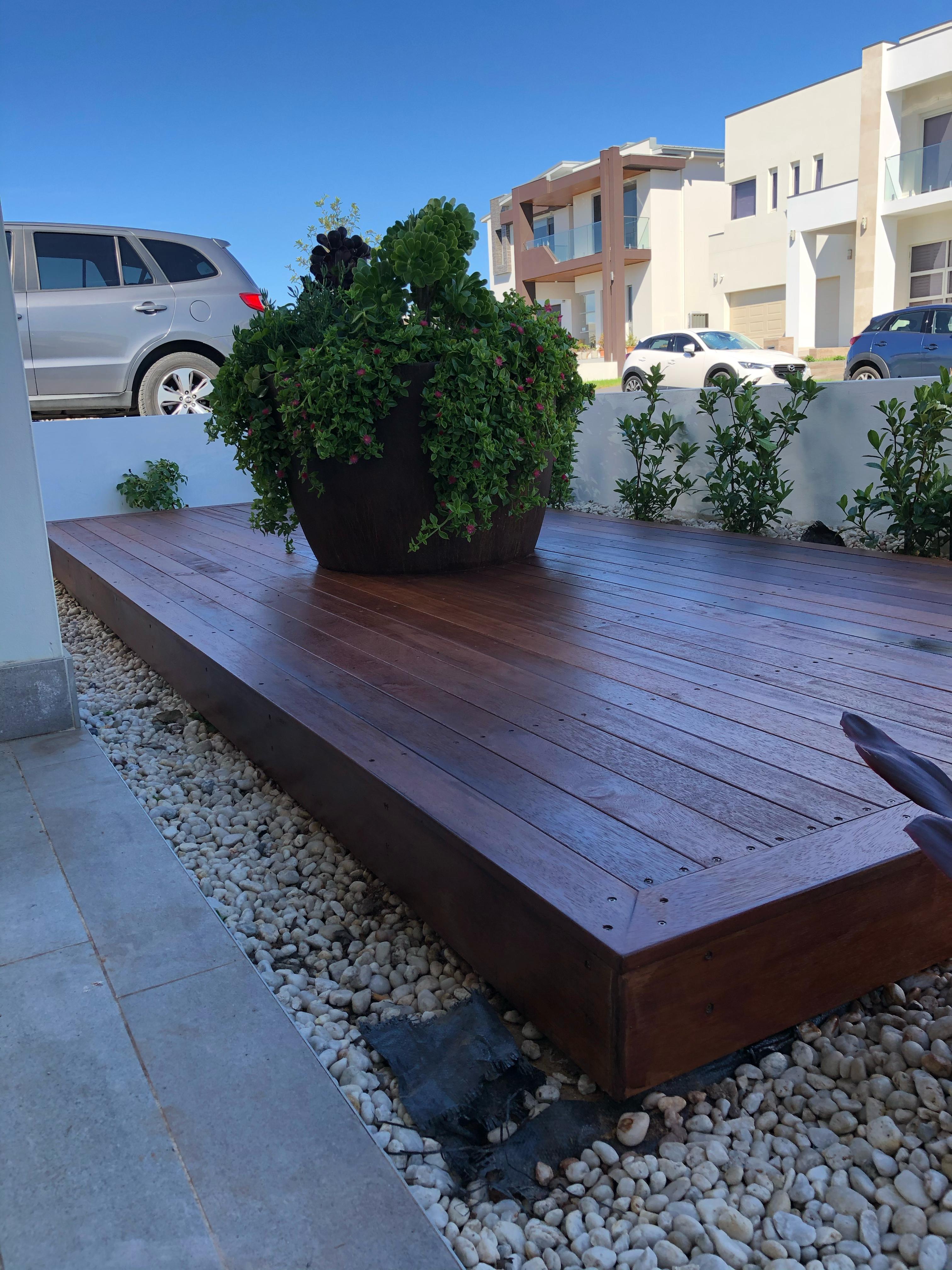 Elevated timber deck with smooth polished finish, featuring decorative planter and modern landscaping with white stone edging.