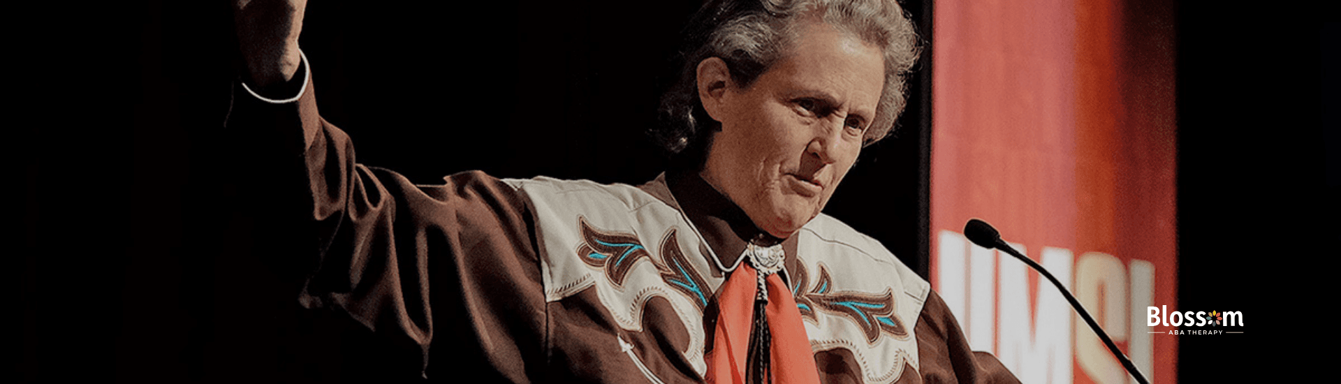 Temple Grandin speaks at a podium, wearing a brown shirt at an autism advocacy event in Georgia.