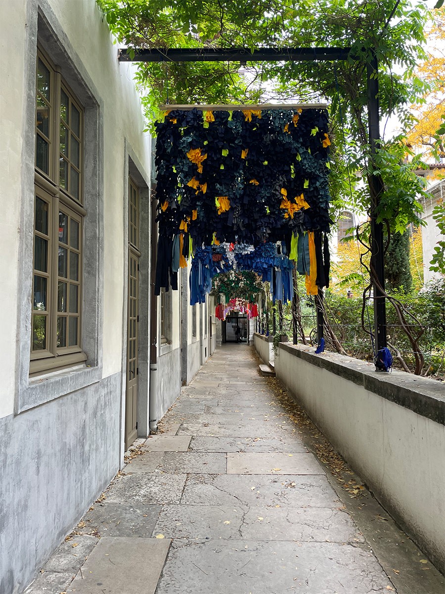 Une installation textile suspendue occupe une allée piétonne extérieure longeant un bâtiment aux murs gris et aux fenêtres encadrées de bois. L'œuvre est composée de multiples bandes de tissus dans des tons dominants de noir et de jaune, suivies d'autres sections aux couleurs variées (bleu, rose, vert) en perspective. Le passage est pavé de dalles de pierre, bordé par un muret sur la droite et une structure métallique ornée de végétation qui surplombe le chemin.